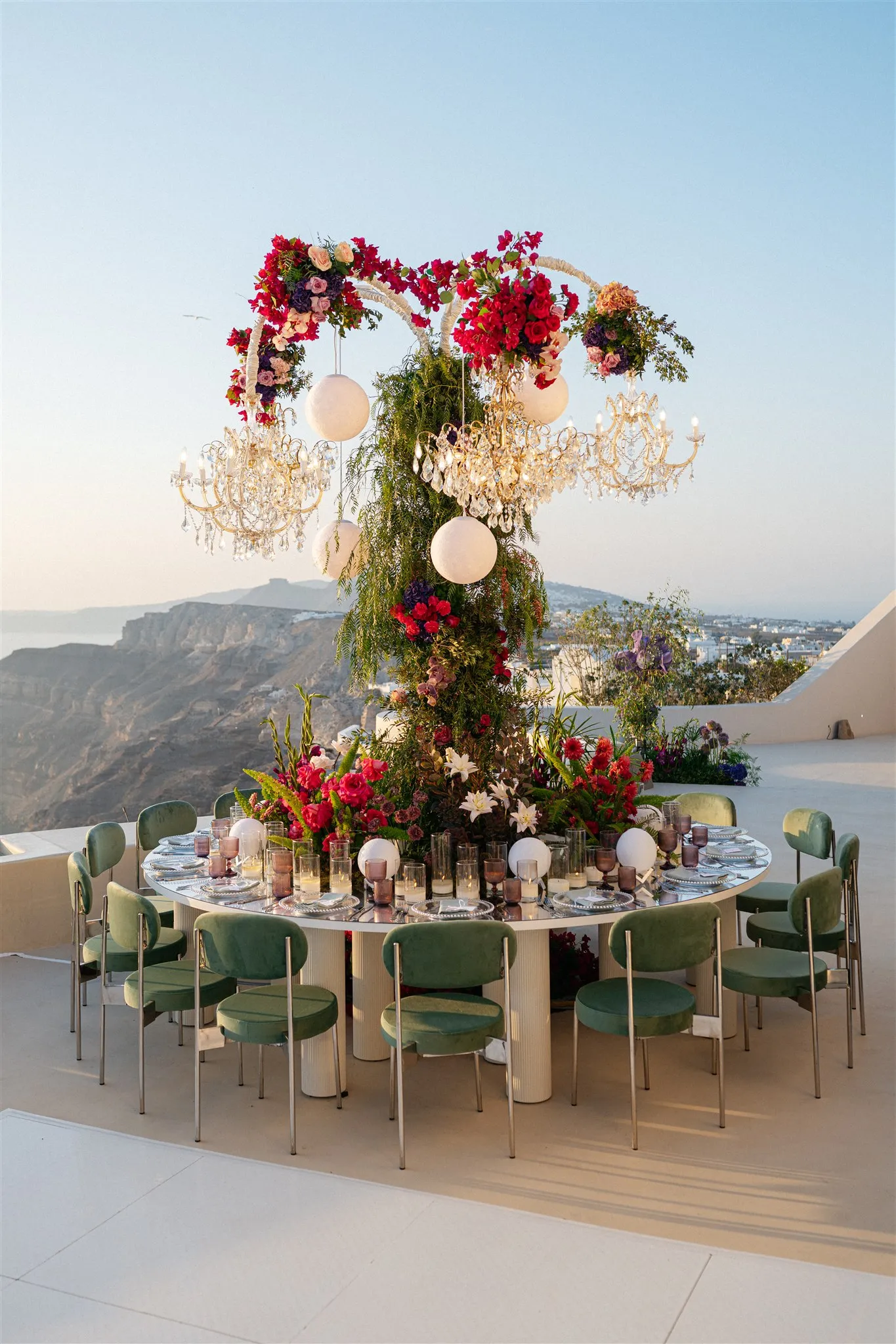 A round white table set for a wedding dinner on a clifftop terrace at Islet Santorini in Thira, Greece, beneath an arch of crimson bougainvillea, red roses and pale peach blooms, with crystal chandeliers and white paper spheres hanging from it. Green velvet chairs surround the table, and a tall column of trailing greenery, white lilies and red flowers rises from its centre, with the caldera cliffs and sea falling away in the soft pink light behind.