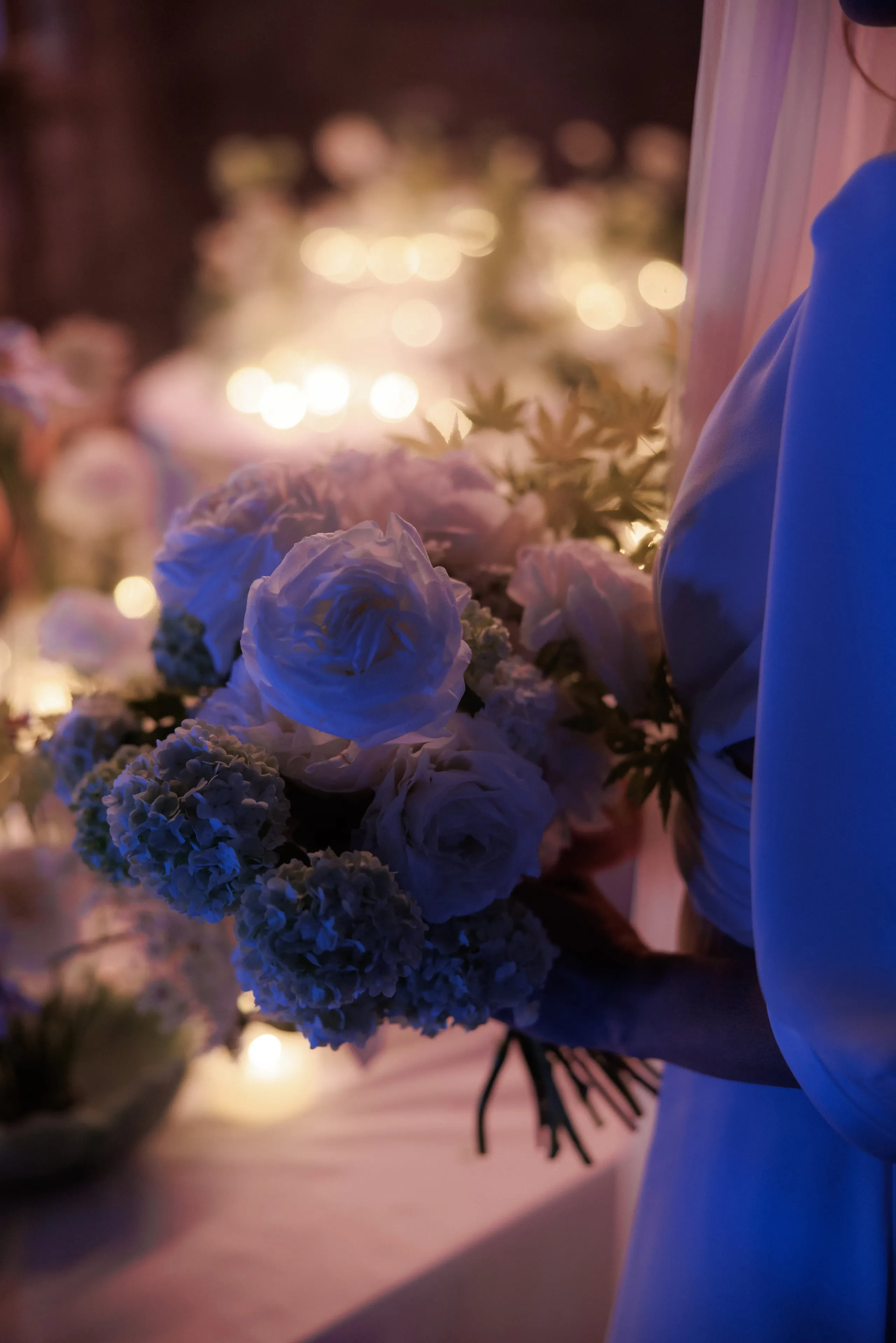 A bridal bouquet held against a blue gown, its pale blue ranunculus and garden roses framed by clusters of blue hydrangea and sprigs of feathery green foliage. Softly blurred string lights glow behind it, and the whole scene is bathed in cool blue light. This wedding flower arrangement was created by manfler in Rethimno, Crete, Greece.