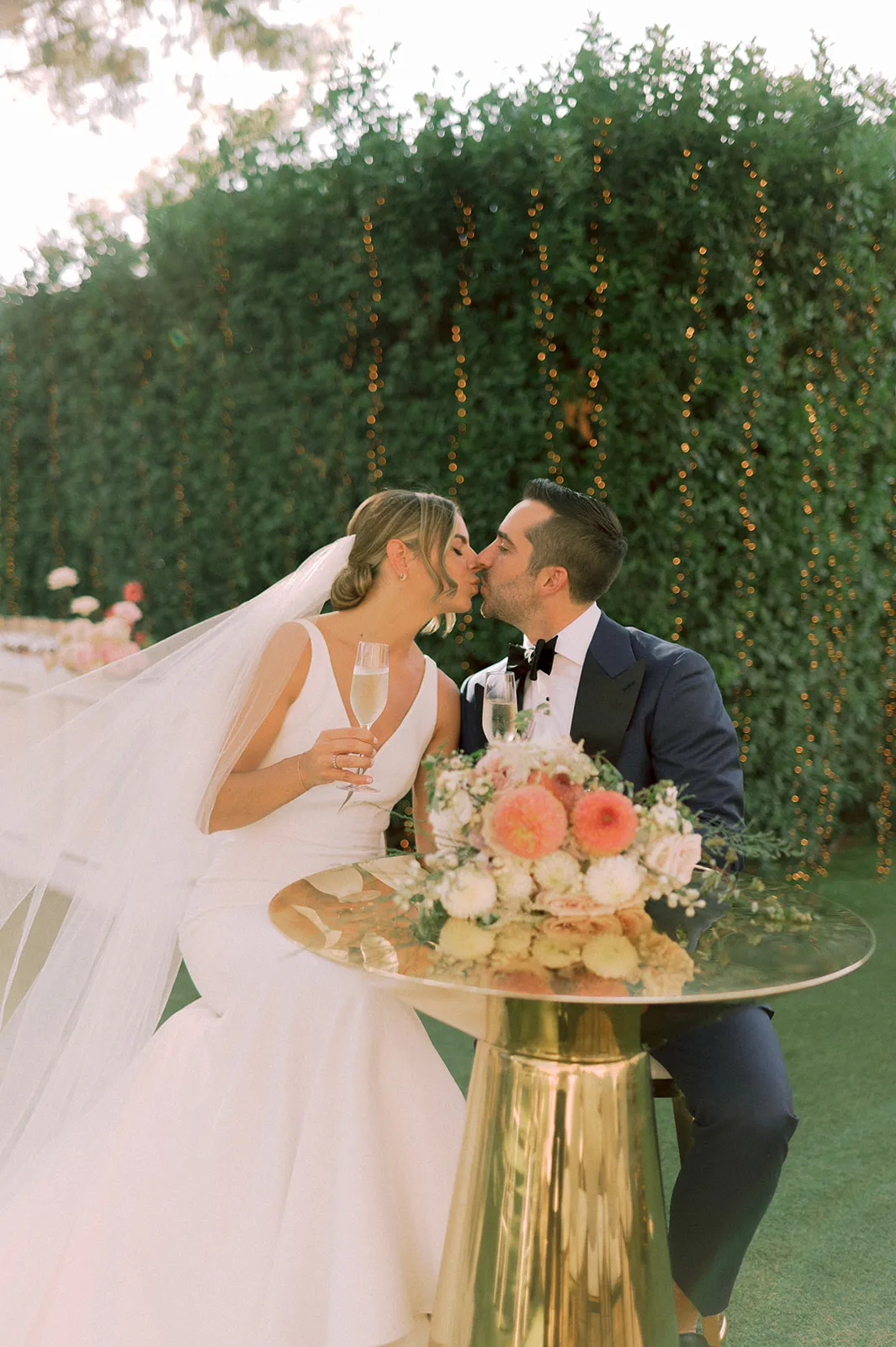 A bride in a sleeveless white gown and a long veil kisses a groom in a navy tuxedo with a black bow tie, both seated at a round mirrored table in front of a dense green hedge strung with warm fairy lights. On the table sits a lush wedding bouquet of peach dahlias, white ranunculus and blush roses, and the bride holds a champagne flute. This wedding table moment is at Lake Vouliagmeni in Greece, and the soft golden light gives it a romantic, celebratory mood.