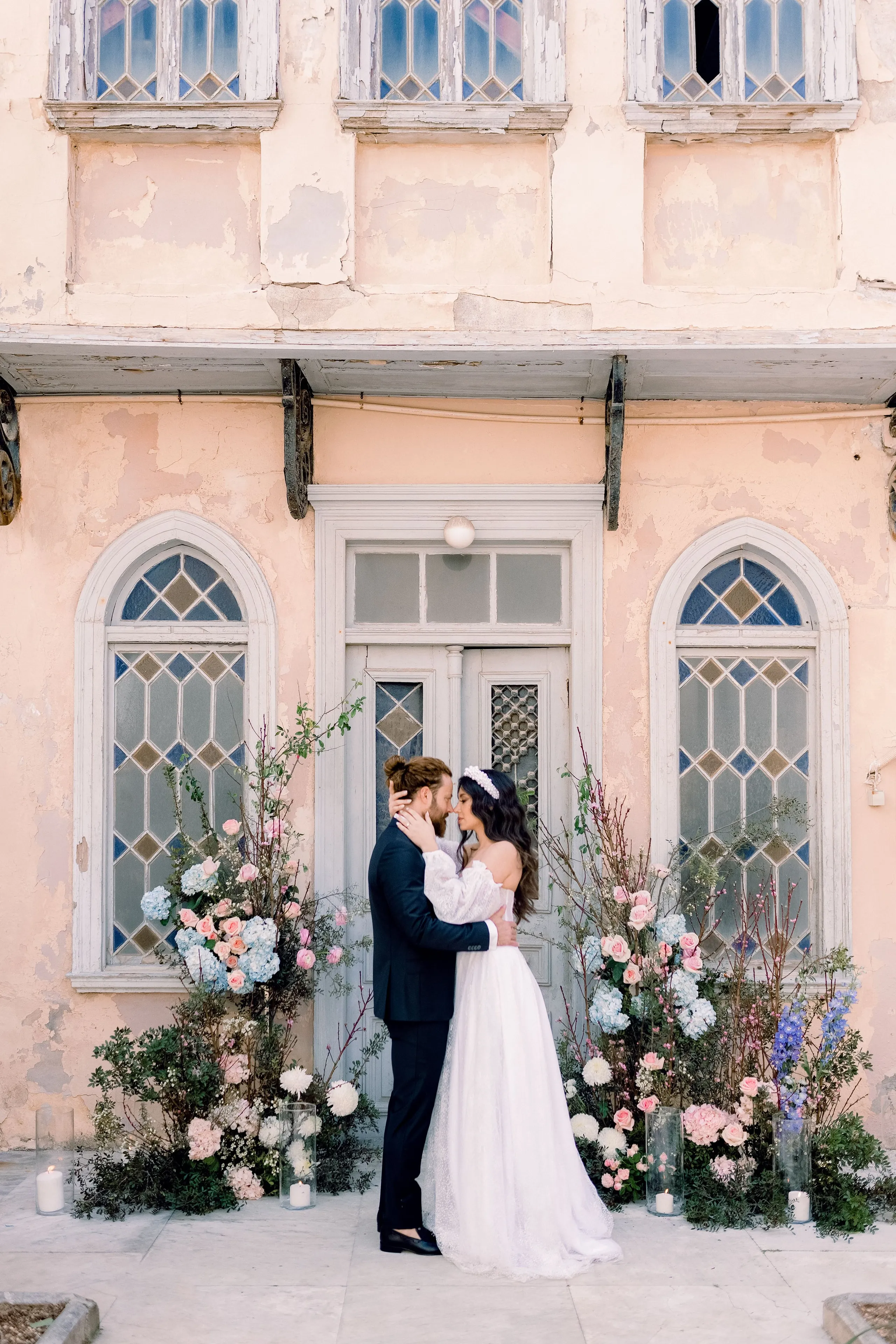 A bride and groom share a tender forehead-to-forehead embrace in front of a pale wooden door in a peach-washed building in Greece, framed by tall floral installations of pink roses, pale blue hydrangeas, white dahlias and delphiniums with candles in glass cylinders at their feet. The groom wears a dark suit and the bride a white gown with a lace-sleeved, off-the-shoulder bodice and a pearl-studded headband, styled as a wedding photograph.
