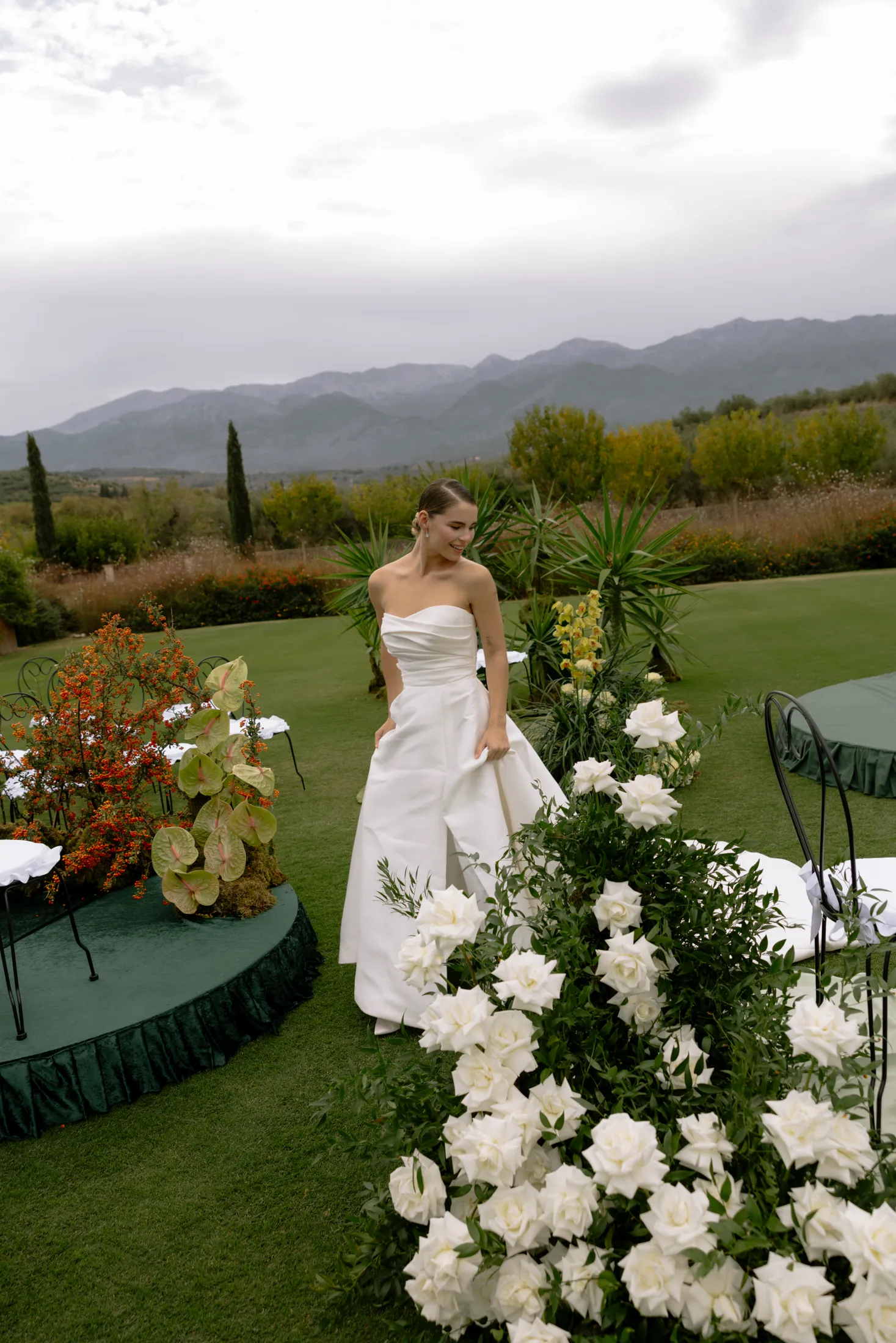 A bride in a strapless white satin gown with a fitted bodice and a gathered skirt walks across a lush green lawn at Ktima Roxani Estate in Crete, Greece, her smile turned to one side. Along the path beside her runs a long line of white garden roses and trailing green foliage, and nearby a round table with a dark green skirt holds a low arrangement of orange berries and chartreuse anthurium. Behind her, a range of misty mountains and soft, cloudy light set a calm, romantic mood.
