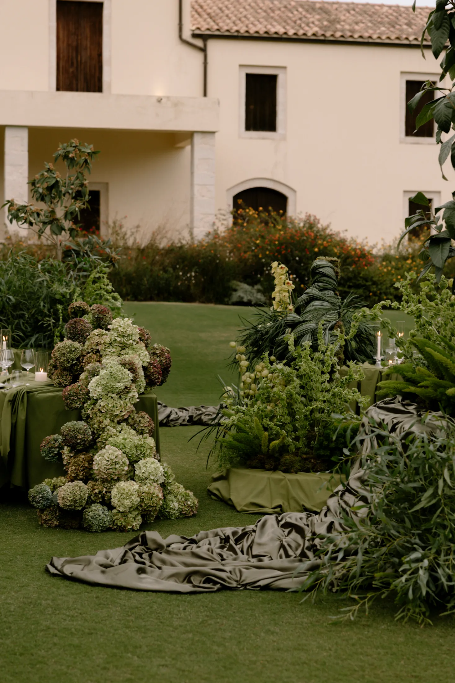 Tiers of green and blush-tinged hydrangeas spill down from a tall mound beside a long table draped in olive-green satin, with more satin pooling across the lawn like spilled silk. Beside it, dark foliage, ferns and tall green flower spikes rise from a low planter, and olive branches arch over the scene. This wedding table styling sits on the grass of Ktima Roxani Estate in Crete, Greece, with a cream stone villa and warm green garden behind.