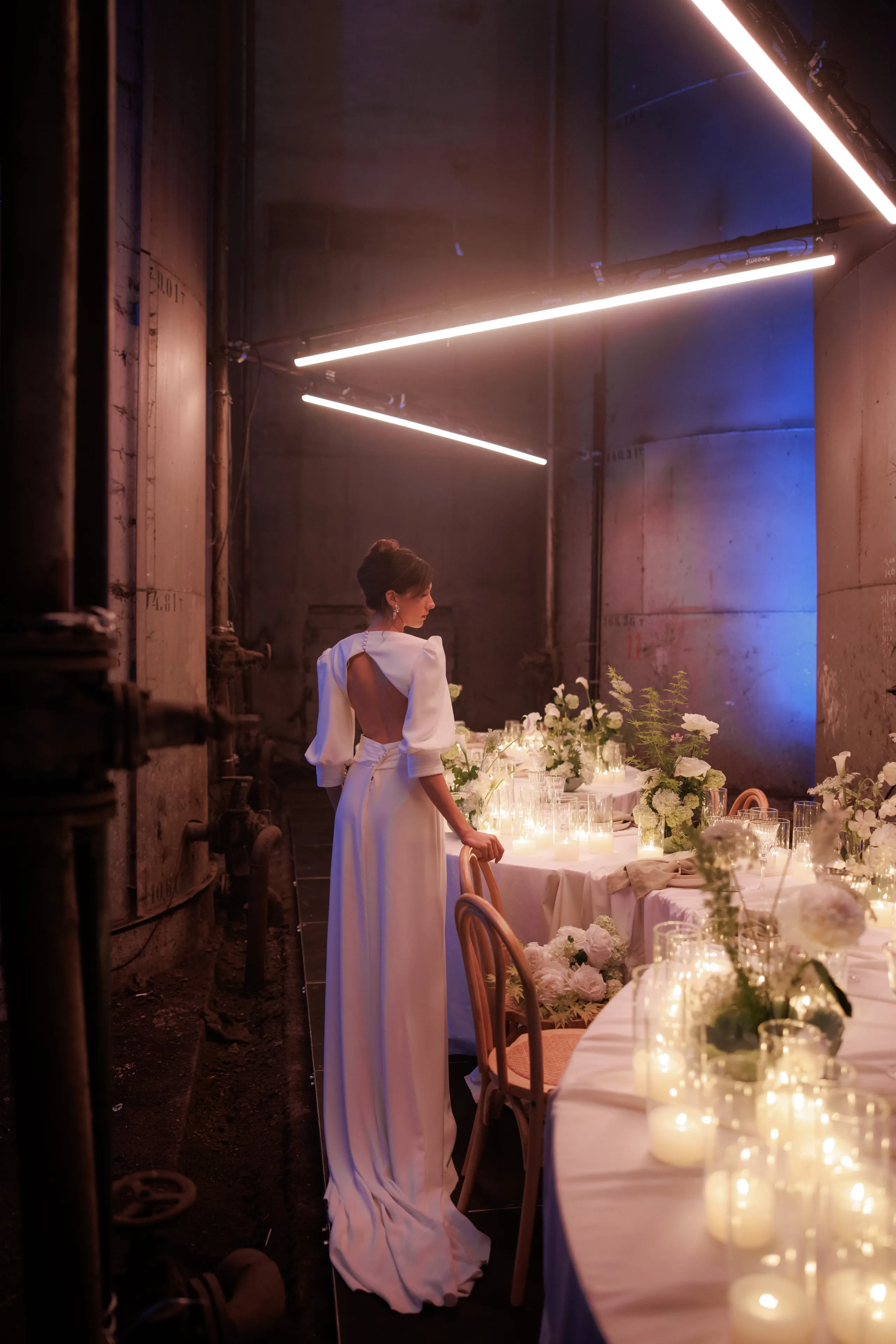 A bride in a long, backless white satin gown with full sleeves stands with her hand resting on the back of a bentwood chair, gazing down a narrow, industrial hall in Rethimno, Crete, Greece. Beside her, a long table is dressed in white linen and set with rows of pillar candles in glass hurricanes and white flowers, including roses and hydrangeas. Overhead, tubes of cool white light and a soft blue wash contrast with the warm candlelight and the weathered concrete walls.