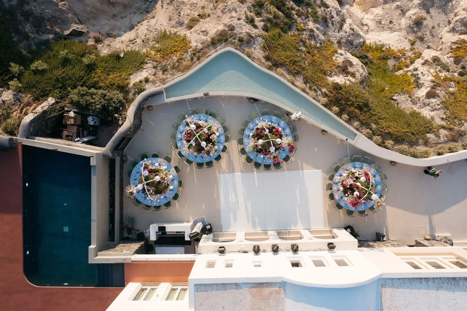 An overhead aerial view of four round wedding tables on a white terrace at Islet Santorini in Thira, Greece, each ringed with pale chairs and topped with low centrepieces of pink and red blooms, greenery and white orbs against blue-rimmed plates. The terrace curves along a cliffside edge of dry scrub and pale rock, with a long rectangular pool to one side and a bar counter in the foreground.