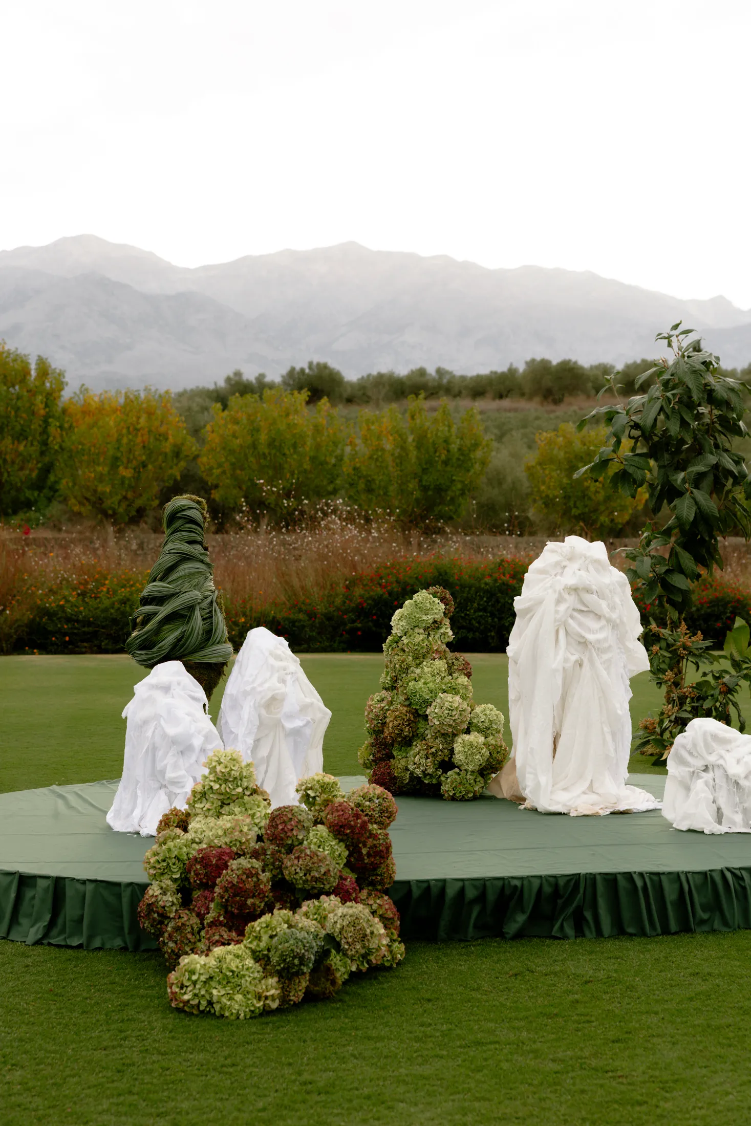 Sculptural wedding installations on a round green stage in the lawn at Ktima Roxani, a wedding venue on Crete, Greece: in the foreground, a heap of hydrangeas in pale lime and dusty burgundy, and behind it a tall cone of hydrangeas in the same greens, while white draped fabric shapes rise like cloaked figures among them. A twisted green leaf column and a leafy branch frame the scene, with misty mountains and olive trees beyond.