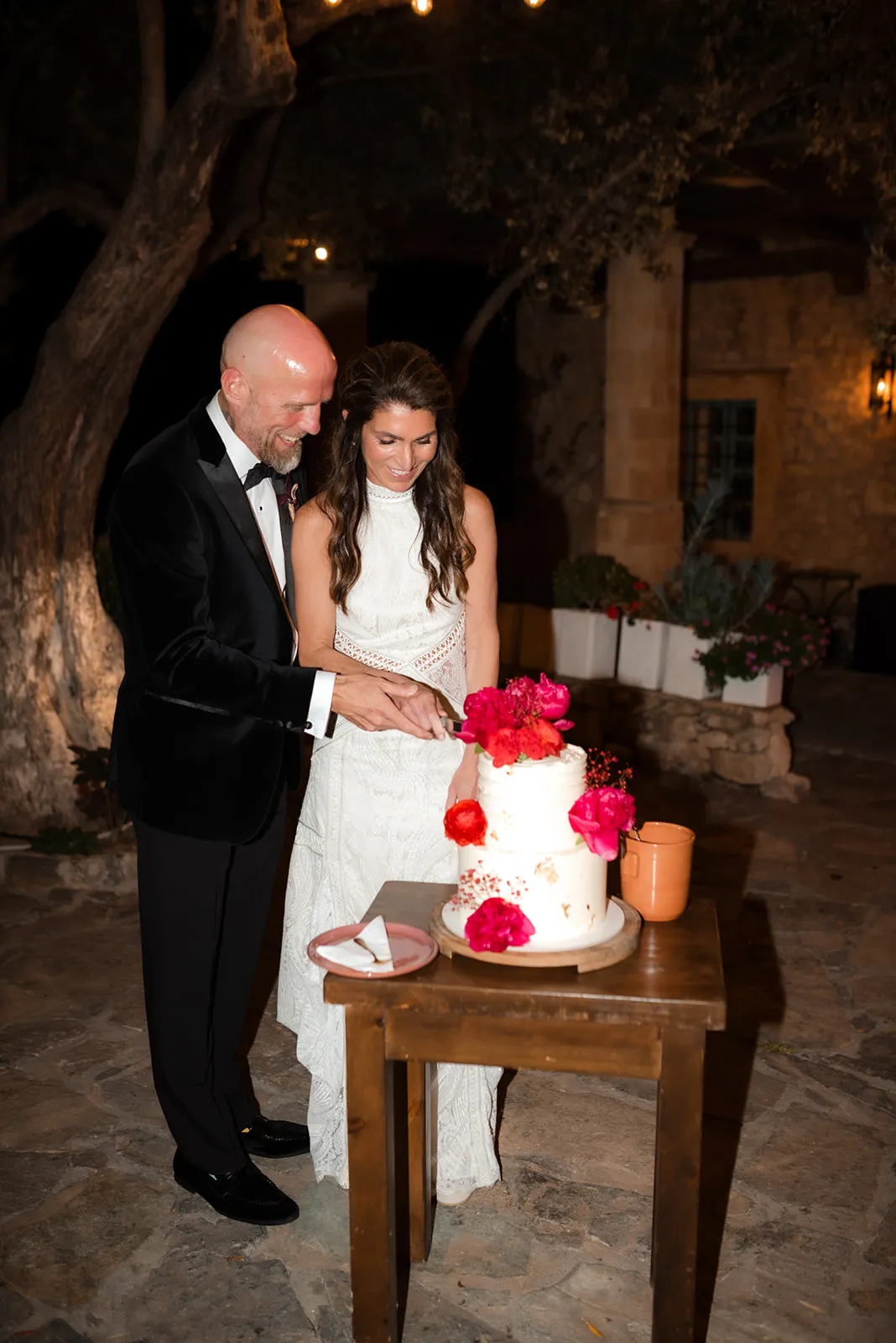 A bride in a fitted white lace gown and a groom in a black tuxedo with a bow tie cut a two-tier white wedding cake together at night, their hands on the knife. Vivid pink and red peonies and small red blooms decorate the cake and its wooden stand, and a pink plate waits on a rustic table. The warm lamplight and stone walls of Agreco Farm in Greece give the scene a cosy, intimate glow.
