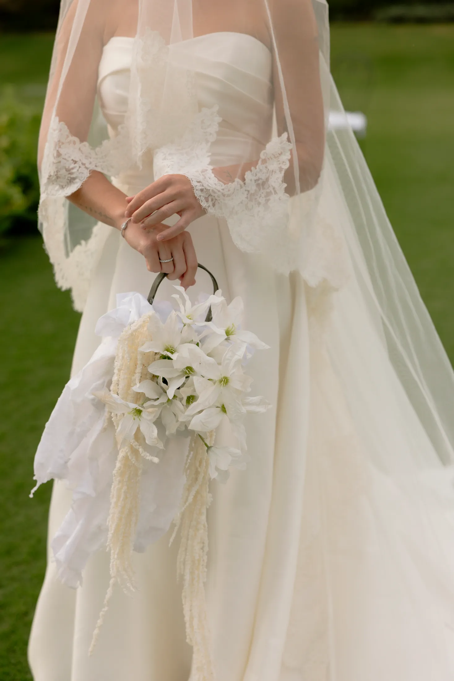 A bride in a strapless satin gown with lace-trimmed sheer sleeves and a long veil stands on lawn, holding a small bouquet by a dark metal ring handle. The bouquet is made of white star-shaped clematis-like blooms with pale green centres, with cream amaranthus tassels and soft white fabric trailing from it. This wedding bouquet was created by manfler for a celebration at Roxani Estate in Armenoi, Crete, Greece.