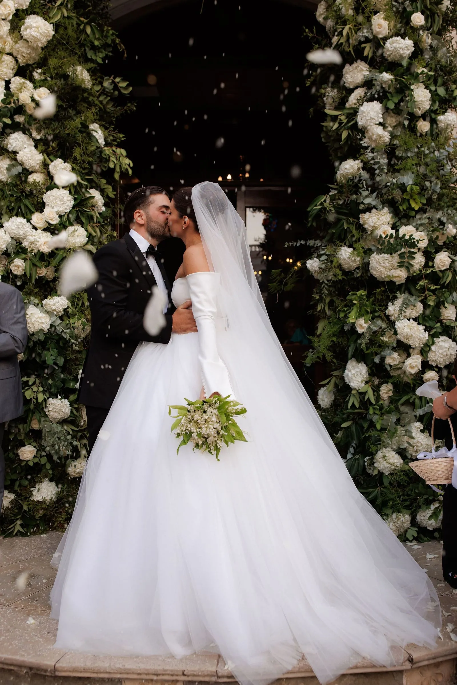 A bride and groom share a kiss beneath a tall doorway framed by lush arches of white hydrangeas, white garden roses and eucalyptus, while white petals drift through the air around them. The bride wears a voluminous white ball gown with off-the-shoulder long sleeves and a flowing veil, and holds a bouquet of lily of the valley wrapped in green foliage. The groom wears a black tuxedo and bow tie. Wedding flowers in Greece, with a warm, glowing interior behind them.