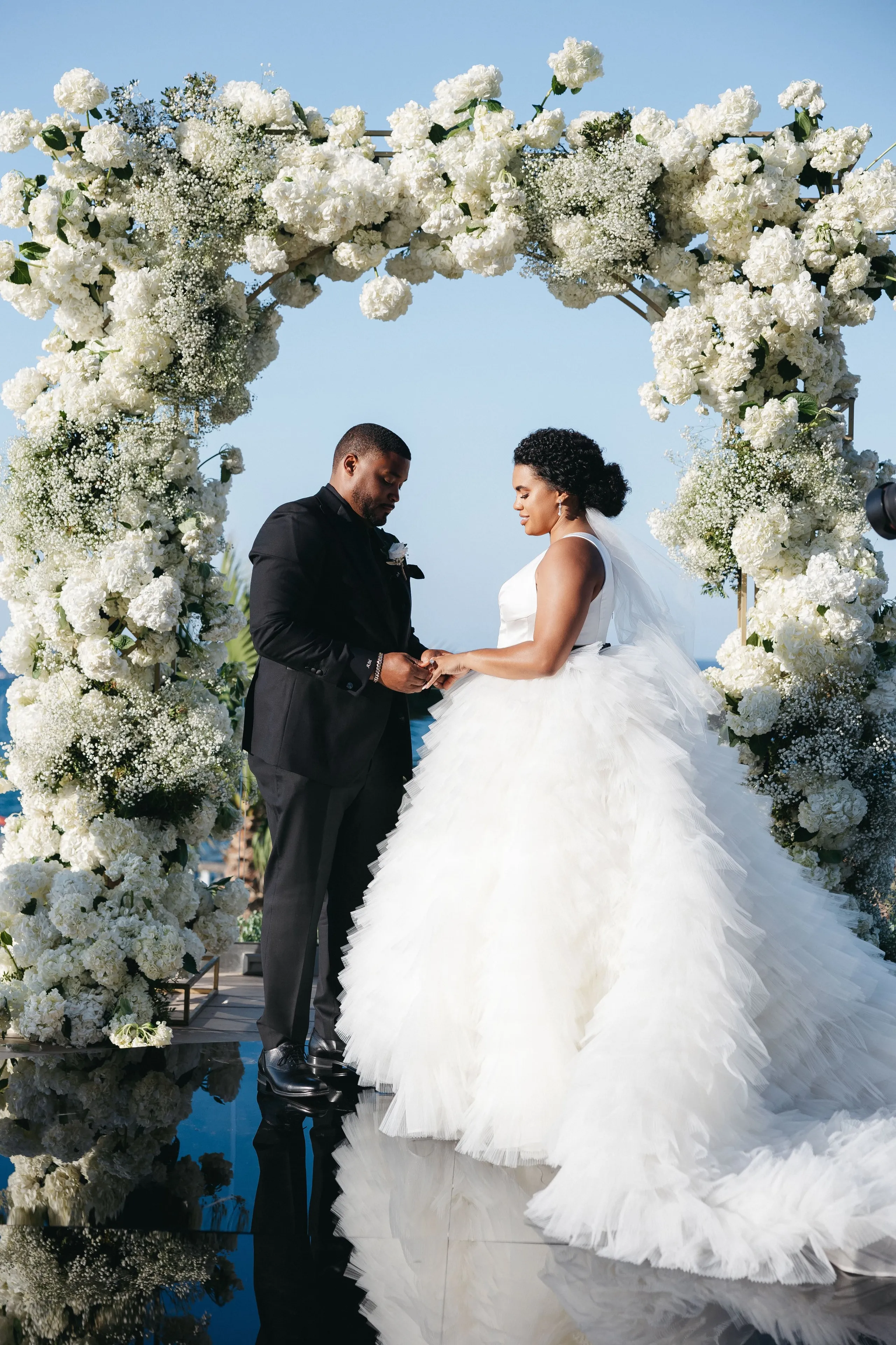 A wedding arch covered in white hydrangeas and baby's breath, set beside the sea at Abaton Island Resort & Spa in Crete, Greece, where a bride in a voluminous tiered tulle gown with a long train stands facing a groom in a black suit as they exchange rings. The bride's hair is swept into a low bun, and the pale blue sky and still water beneath the arch reflect the white blooms in a soft, airy scene.