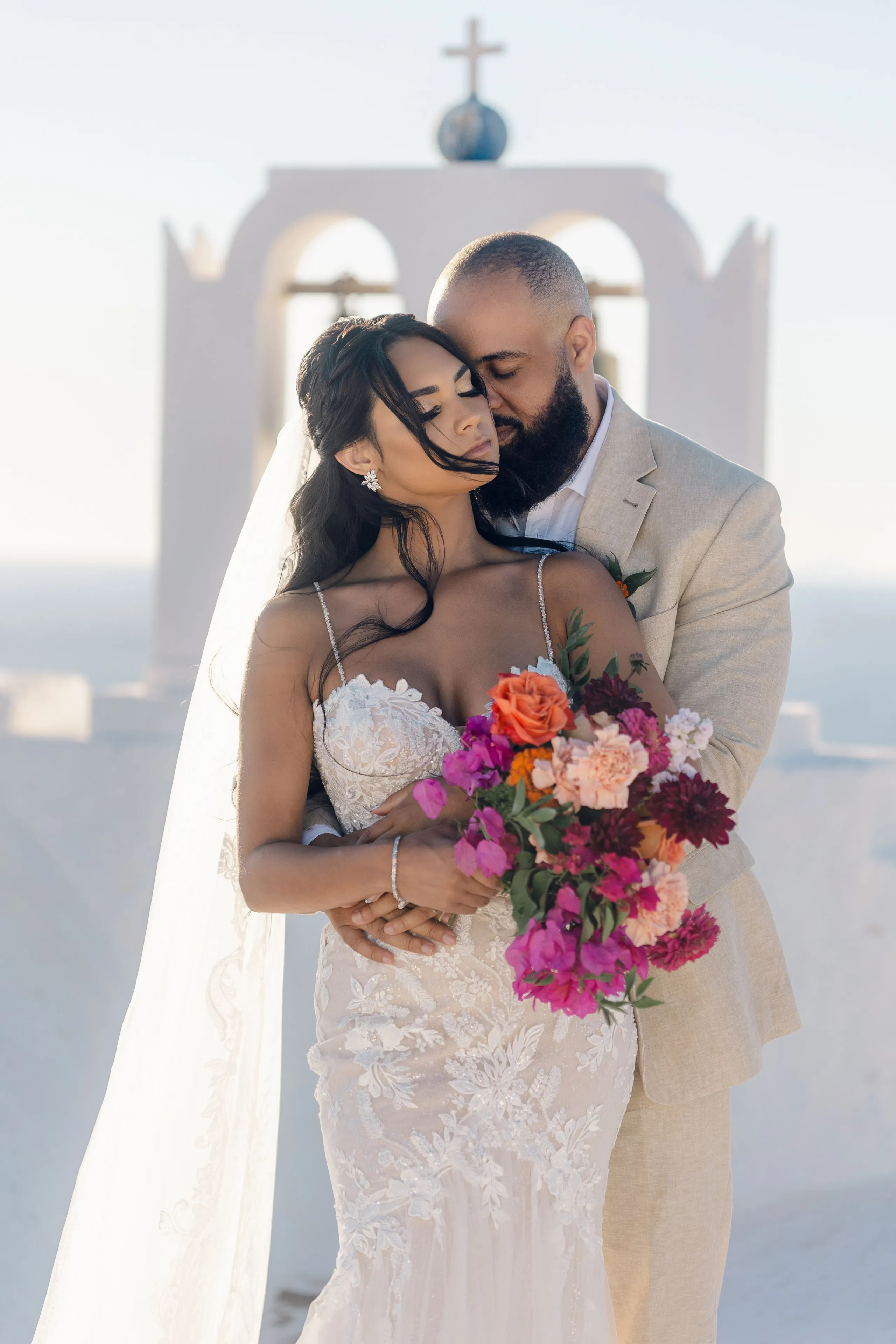 A bride in a lace fitted gown and a long veil stands in the embrace of her groom in a pale linen suit, his head bowed close to hers, both with eyes closed. She holds a bridal bouquet of magenta bougainvillea, deep burgundy dahlias, orange roses and blush garden roses with green foliage. Behind them rises a whitewashed bell tower topped with a cross against a soft, hazy sky, at Cavo Ventus Luxury Villa in Santorini, Greece.