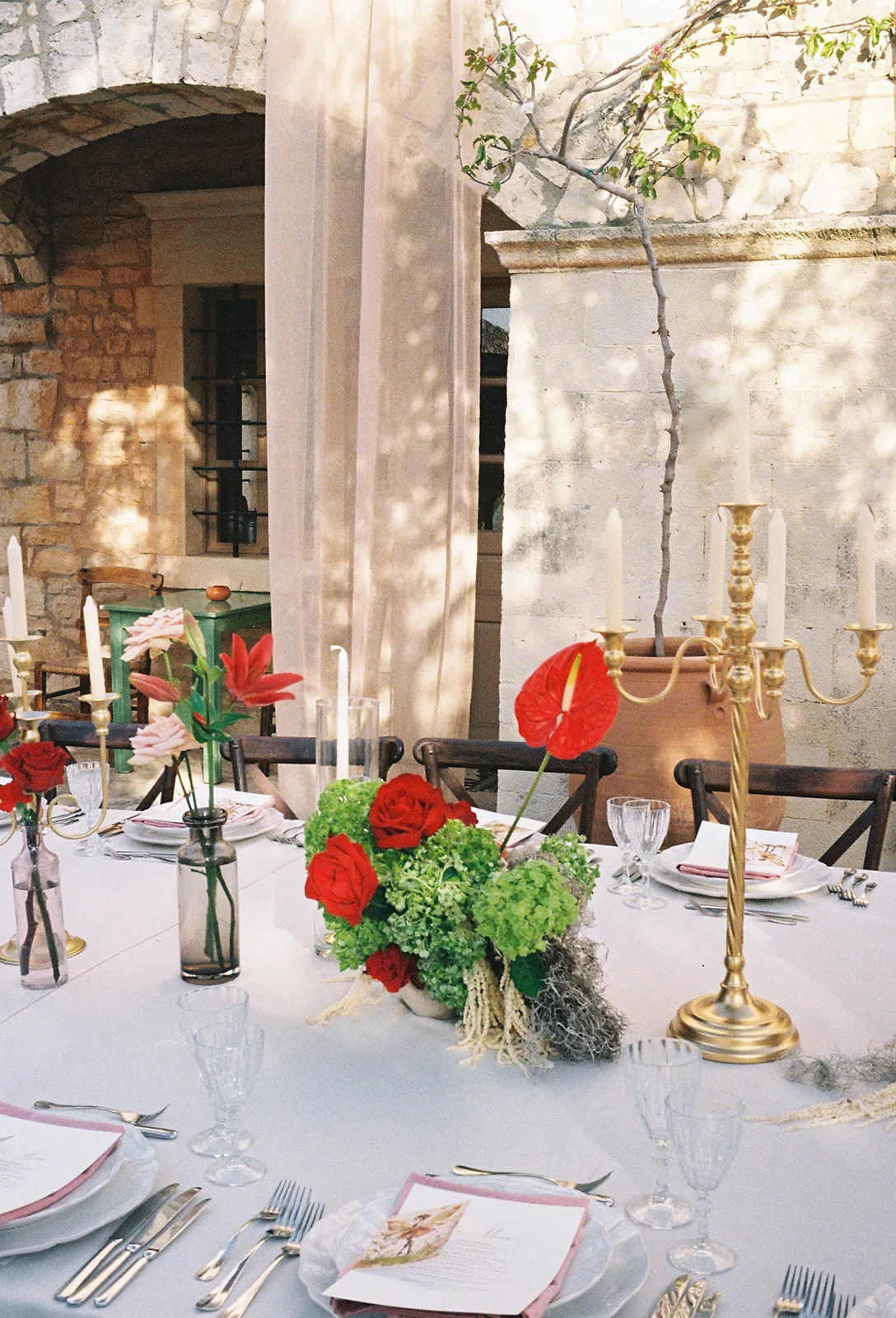 A wedding table set on a sunlit stone terrace at Agreco Farm in Greece, dressed in a centrepiece of red garden roses and green hydrangea with trailing moss, plus a single red anthurium rising from the arrangement. Slim glass bud vases hold pink roses and red lilies, and tall gold candelabras with white tapers stand among the place settings. Soft sheer curtains and a terracotta urn frame the rustic scene in warm, dappled light.