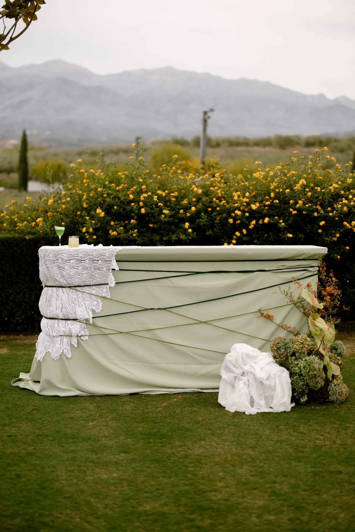 A sage-green draped bar table on a lawn, wrapped with dark green cord and layered with white crocheted lace at one end, with a green cocktail glass set on top. Beside it stands an arrangement of pale green hydrangeas, anthurium leaves and russet twigs, and behind it a hedge of yellow flowers below grey mountains. This is an event flower and styling display at Ktima Roxani Estate in Armenoi, Crete, Greece.