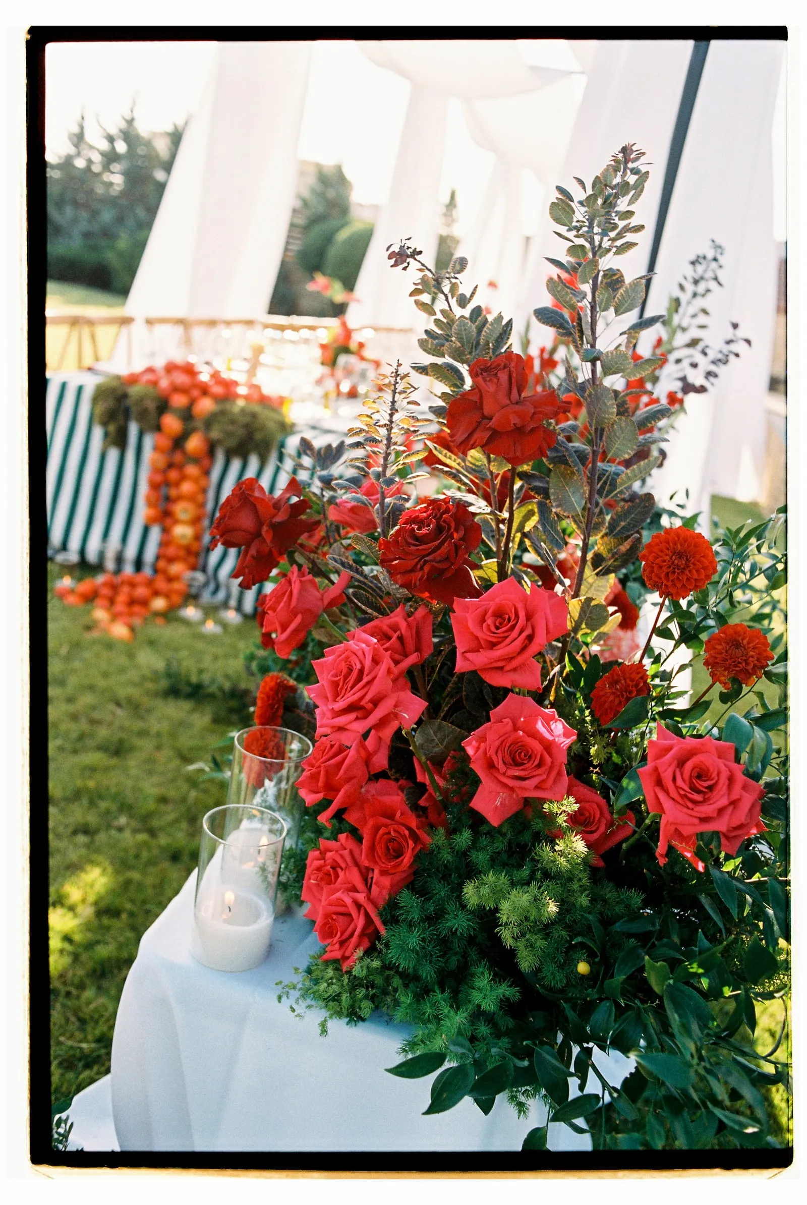 A large, lush arrangement of red garden roses in shades from crimson to coral, paired with orange dahlias and sprigs of dark-leaved eucalyptus and soft green foliage, sits on a white-draped table on the grass. Two glass hurricane candles glow at its base, and behind it a striped table is lined with orange fruit and more blooms beneath white fabric canopies. This wedding flower display is set in a garden ceremony at Wine Dark Sea Villas in Santorini Villa, Rodia, Crete, Greece.