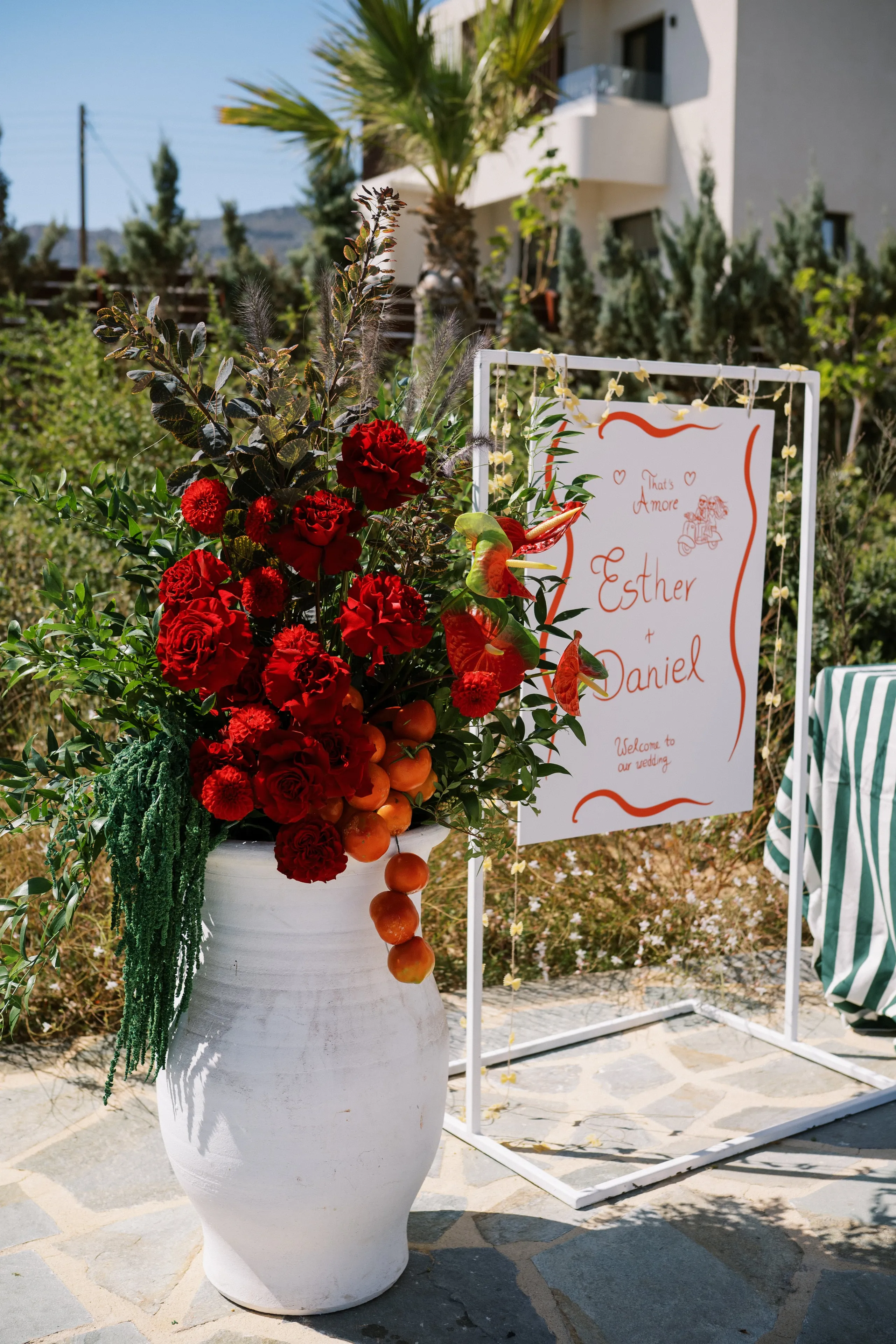 Red garden roses and carnations spill from a tall white ceramic urn on a stone terrace, with trailing green amaranthus, dark eucalyptus, and clusters of small orange kumquats hanging down its side. Beside it stands a white welcome sign with red script reading "That's Amore, Esther & Daniel, Welcome to our wedding," framed by red anthurium blooms and tiny white flowers strung along its edge. This wedding display at Wine Dark Sea Villas on Crete, Greece, sits under bright daylight with pines and a palm behind it.