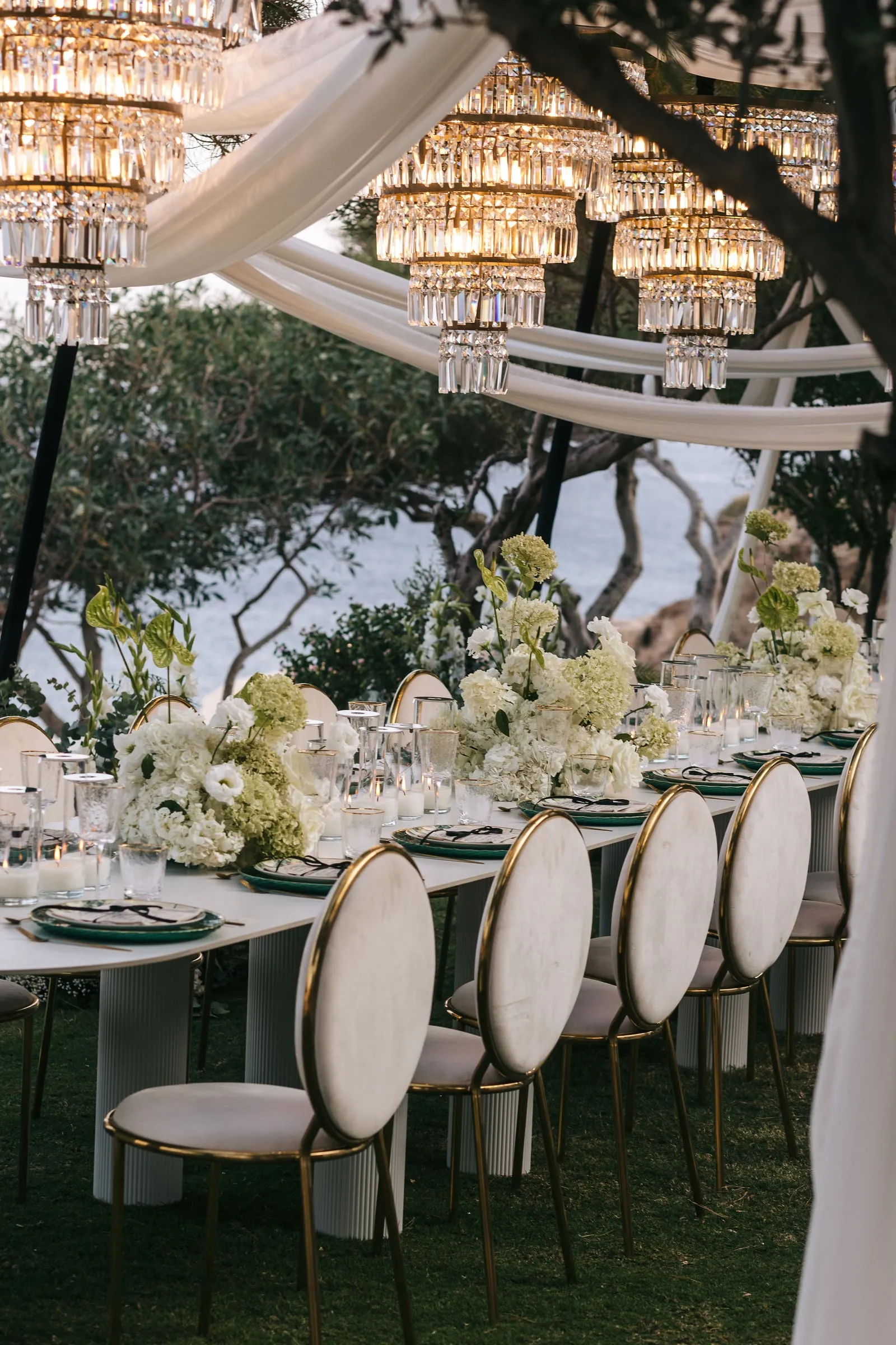 A long wedding dining table on a grassy terrace under a canopy of draped white fabric and tiered crystal chandeliers, with the sea glimpsed through olive trees beyond. Low centrepieces of white and pale green hydrangeas, white roses and lily-like blooms with green foliage run down the table, flanked by cream high-backed chairs with gold frames and glassware set on green-rimmed plates. This wedding table setting is at Cycladic Gem Luxury Villa on Ios Island, Santorini, Greece.