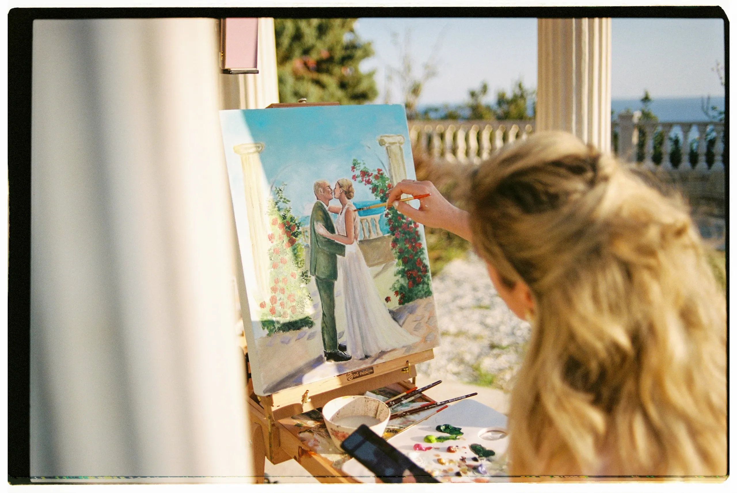 A woman with long wavy blonde hair, seen from behind, paints on a small easel on a sunlit terrace, holding a fine brush to a canvas. The canvas shows a couple in a tender kiss beneath white columns and red climbing flowers, with the sea beyond. This is a wedding illustration painted live at Wine Dark Sea Villas in Crete, Greece, with palettes, brushes and a water cup on the table beside her.