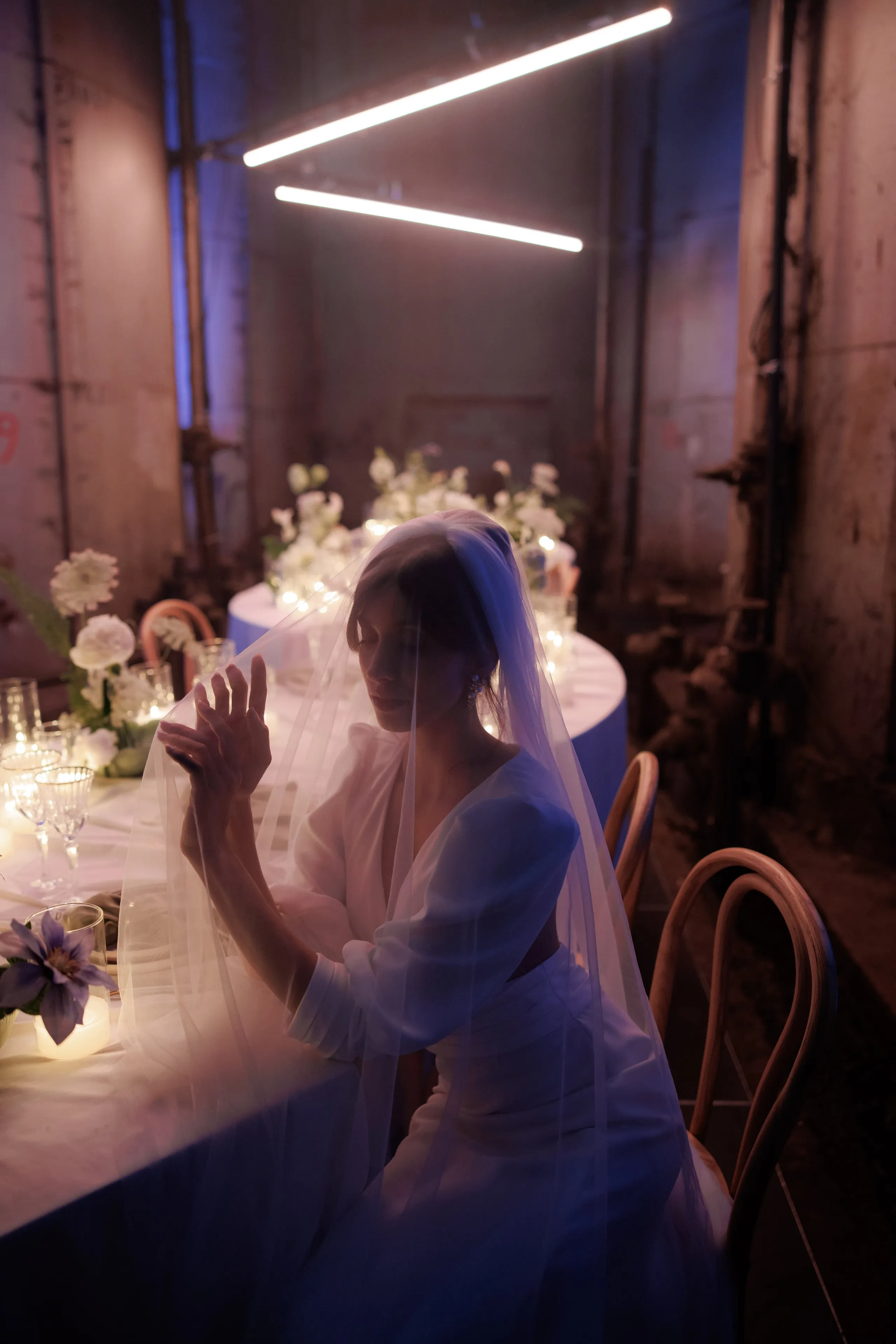 A bride seated at a long table in a dim, industrial room in Rethimno, Crete, Greece, her face softened by a sheer veil that drapes over her shoulders and across the table. Her hands are raised together in front of her, lit in violet and blue from the neon tubes overhead. Around her, white dahlias and sprays of white blooms rise from candlelit table arrangements, with a trailing purple clematis bloom in the lower left, all part of the wedding table flowers in Greece.