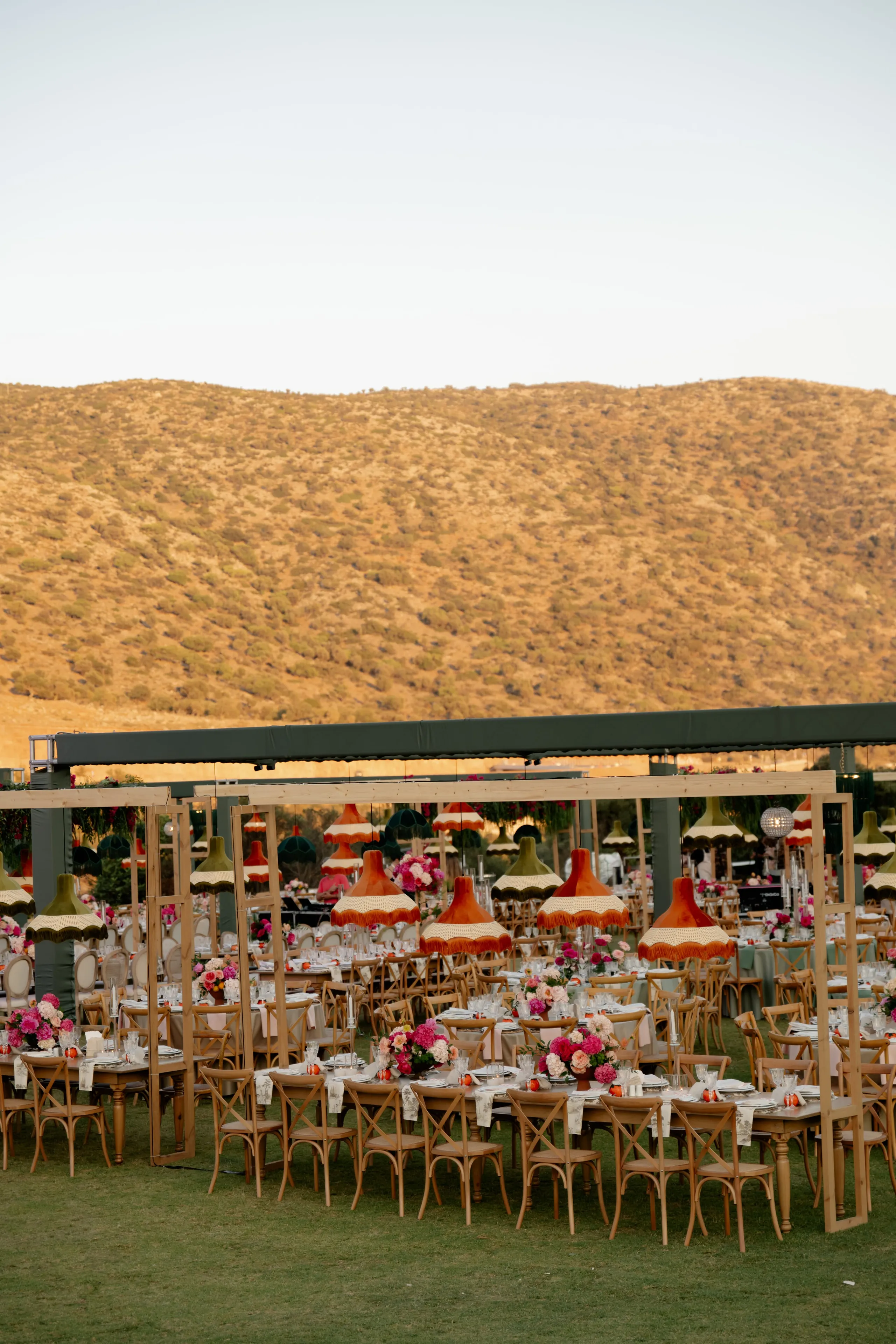 Long rows of wooden cross-back chairs line white-clothed tables on a green lawn, each centred with pink and coral blooms in low arrangements, possibly garden roses and peonies. Above them hang rust-orange and olive-green fringed lampshades under a dark green canopy, with a sun-baked golden hillside behind. This wedding reception at Crete Golf Club in Chersonisos, Greece, is lit by the warm glow of early evening.