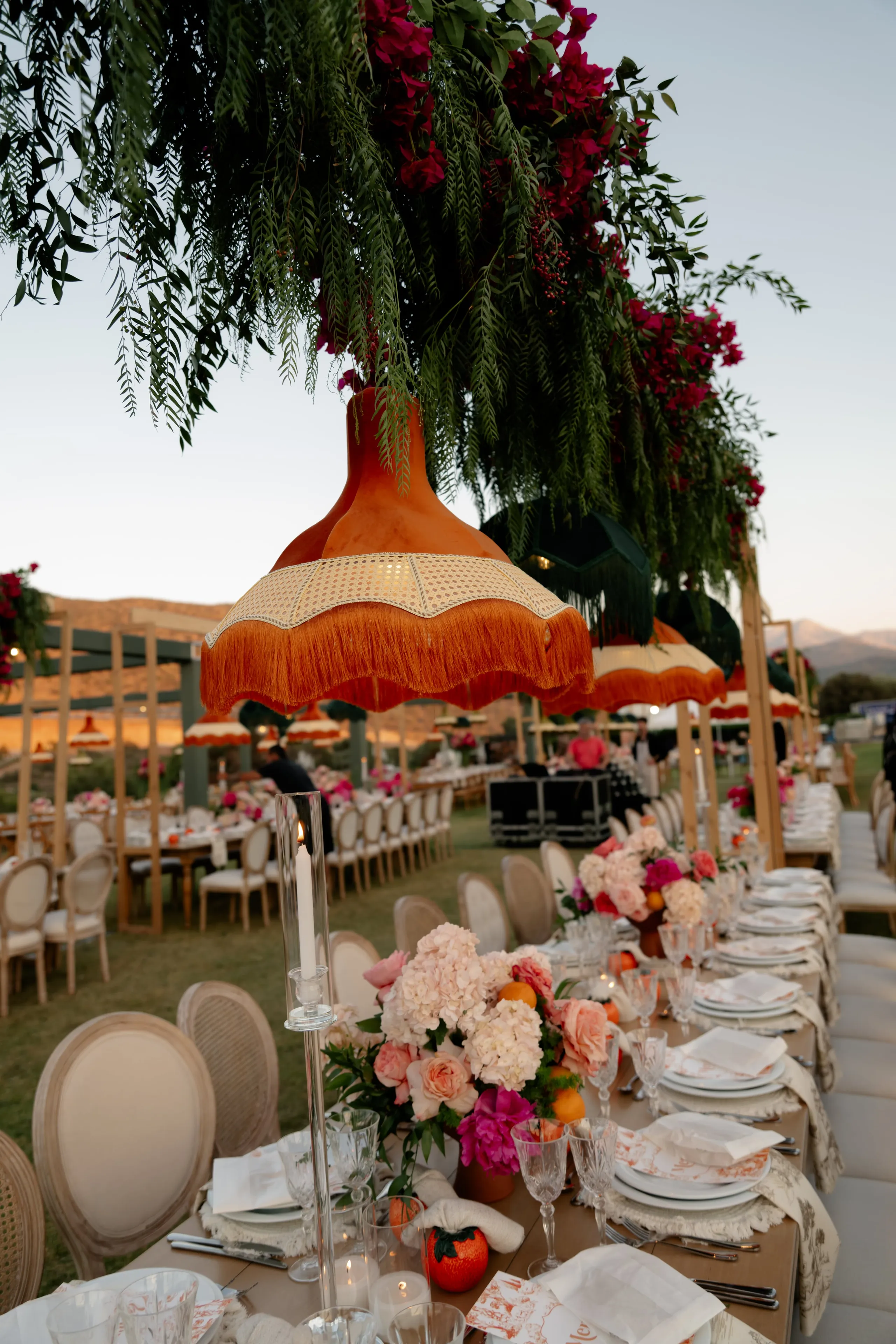 A long outdoor wedding table at Crete Golf Club in Chersonisos, Greece, laid for an evening dinner beneath hanging orange fringed lampshades and trailing green foliage with magenta bougainvillea. The centrepiece of peach garden roses, pale hydrangeas and pink blooms sits among small oranges, with clear glass candle holders, white plates and linen napkins along the table. Golden-hour light fades over the hills behind rows of cane-backed chairs.