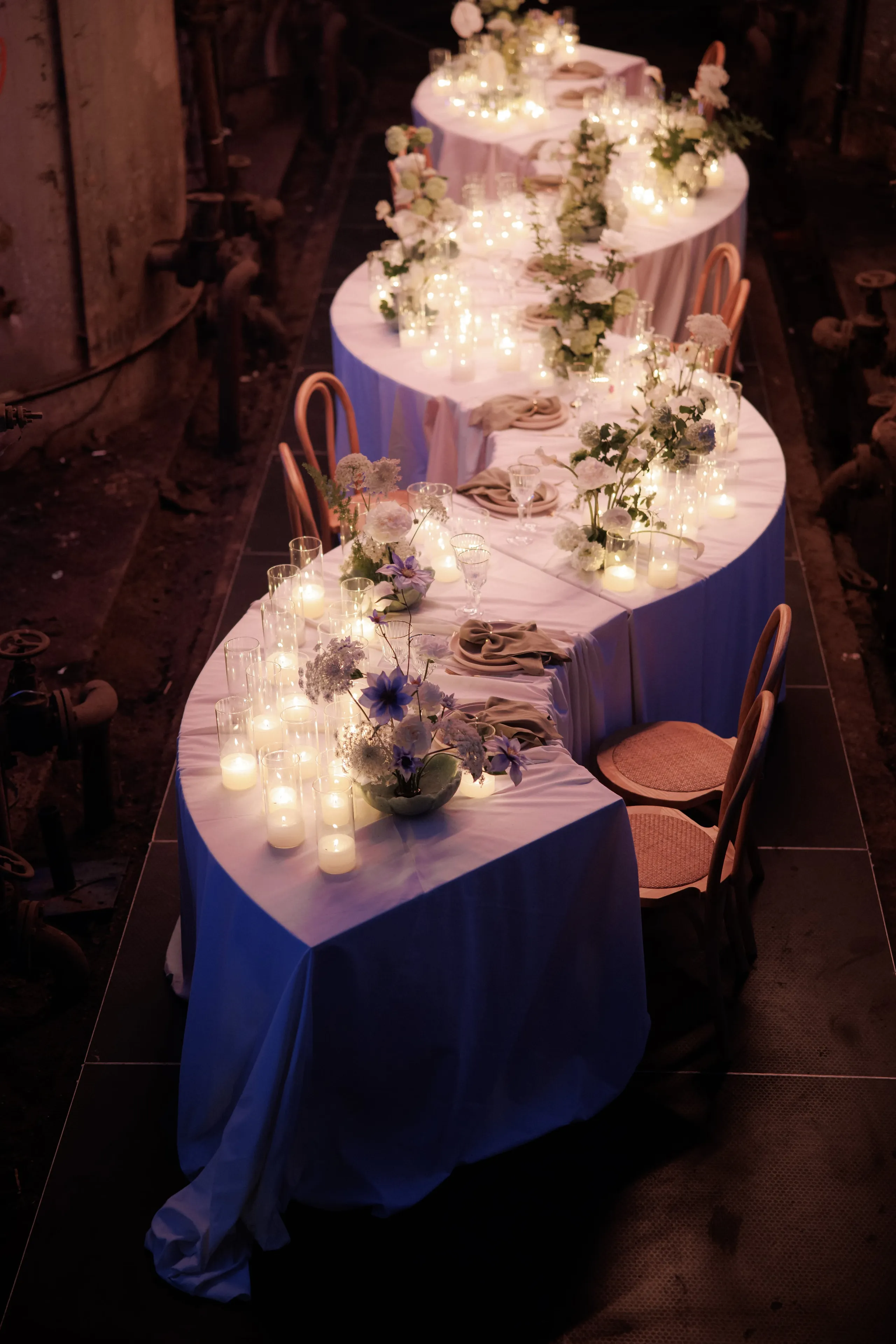 A long, curving banquet of tables draped in soft lavender-blue linen, lit by rows of pillar and tealight candles in glass holders. Low arrangements of white hydrangea, violet clematis and pale blue scabiosa sit between taupe napkins, with bentwood chairs with woven seats lining the outside. This wedding table setting in Rethimno, Crete, Greece, is set in a dim industrial space with pipes along the walls, and the candlelight makes the whole scene feel intimate and warm.