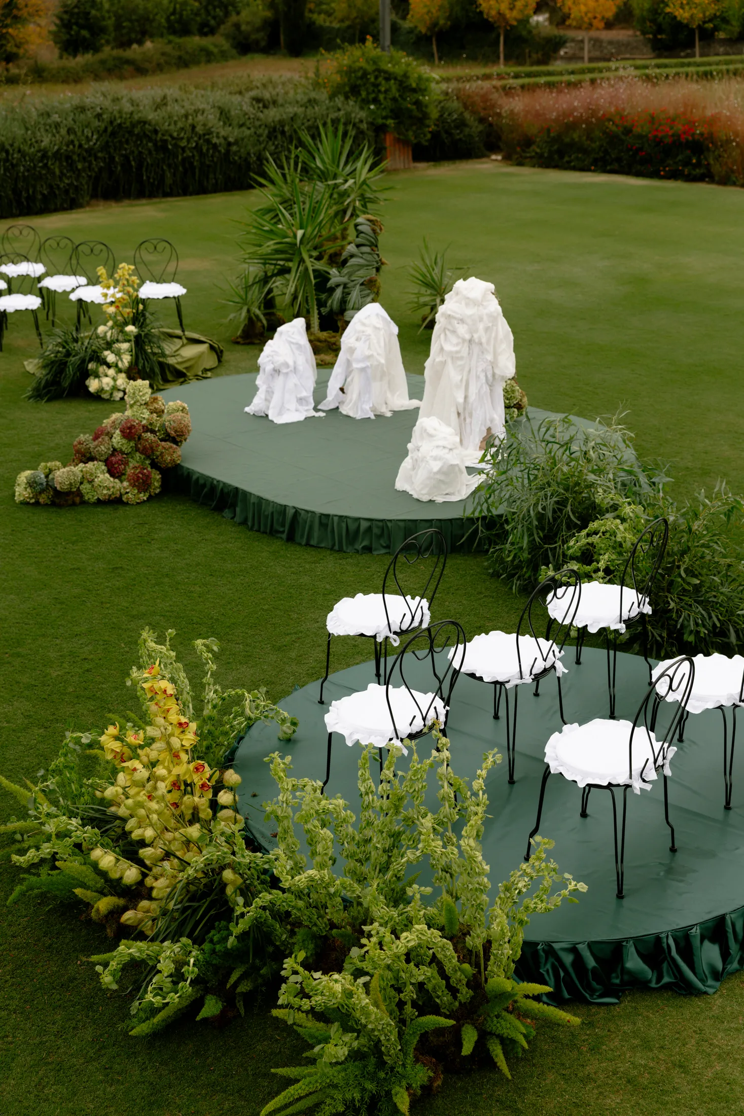 Elevated view of a wedding garden set on a lawn at Ktima Roxani Estate in Armenoi, Crete, Greece, with two round green-draped platforms holding black wrought-iron bistro chairs with white ruffled seat covers. On the lower platform, billowing green foliage and sprays of pale yellow cymbidium orchids frame the chairs, while on the upper platform four tall white draped forms stand among spiky palms and feathery grasses. Hydrangeas in cream and deep burgundy sit at the left edge of the lawn, all under soft, even daylight.