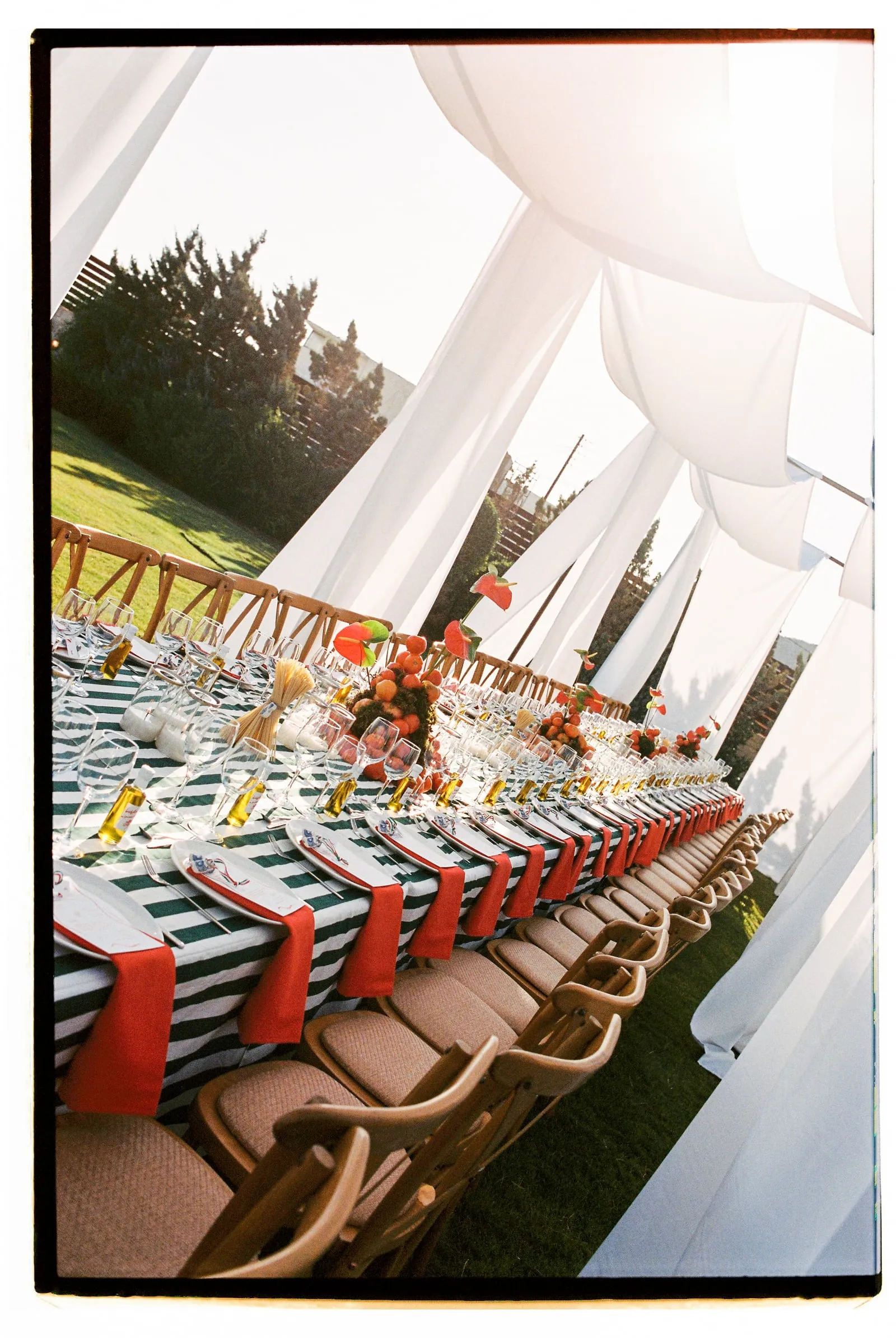 A long banquet table draped in green and white striped linen runs beneath billowing white fabric canopies in a garden at Wine Dark Sea Villas in Rodia, Crete, Greece, set for a wedding reception. Red napkins are folded across each place setting, with clear glassware, gold-tinted stems and cutlery laid out in rows, and centrepieces of orange blooms, red anthuriums and fruit rise along the table. Wooden folding chairs with cushioned seats line both sides, and the soft evening light glows through the fabric onto the green lawn.