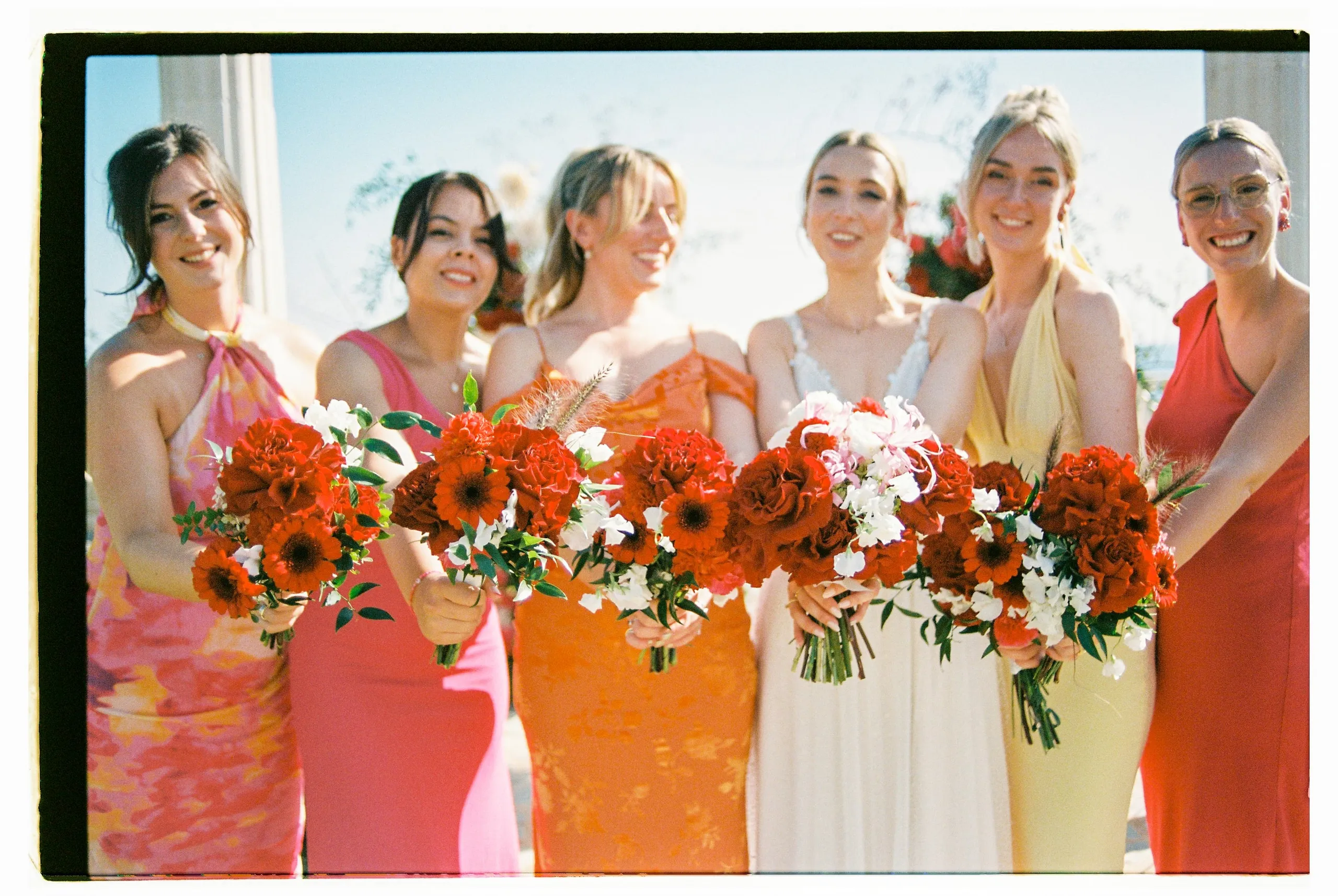 Six women stand side by side holding bouquets of red flowers, with the bride in the centre wearing a white lace-strapped gown and holding a bouquet of white sweet peas and pale pink blooms. The other five, in dresses of pink, coral, orange and yellow, hold red gerbera daisies, deep red garden roses and carnations, accented with white blooms and green leaves. These wedding bouquets were arranged for a garden ceremony at Wine Dark Sea Villas on Crete, Greece, under a bright blue sky.
