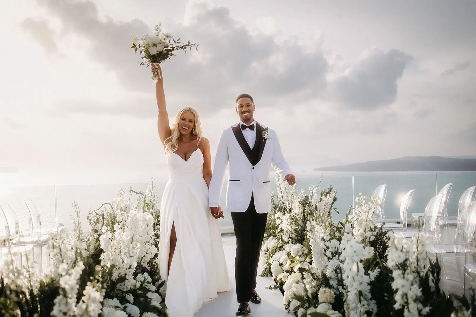 A bride in a white satin gown with a deep side slit raises a white rose and foliage bouquet high in one hand, while her groom in a white dinner jacket with black lapels holds her other hand and smiles beside her. They walk down a white aisle lined with tall white delphinium and stock-like blooms and dark greenery, with rows of clear acrylic chairs beyond. This wedding aisle on a clifftop terrace at Cavo Ventus Luxury Villa in Santorini, Greece, opens onto a soft grey sky and the calm sea.