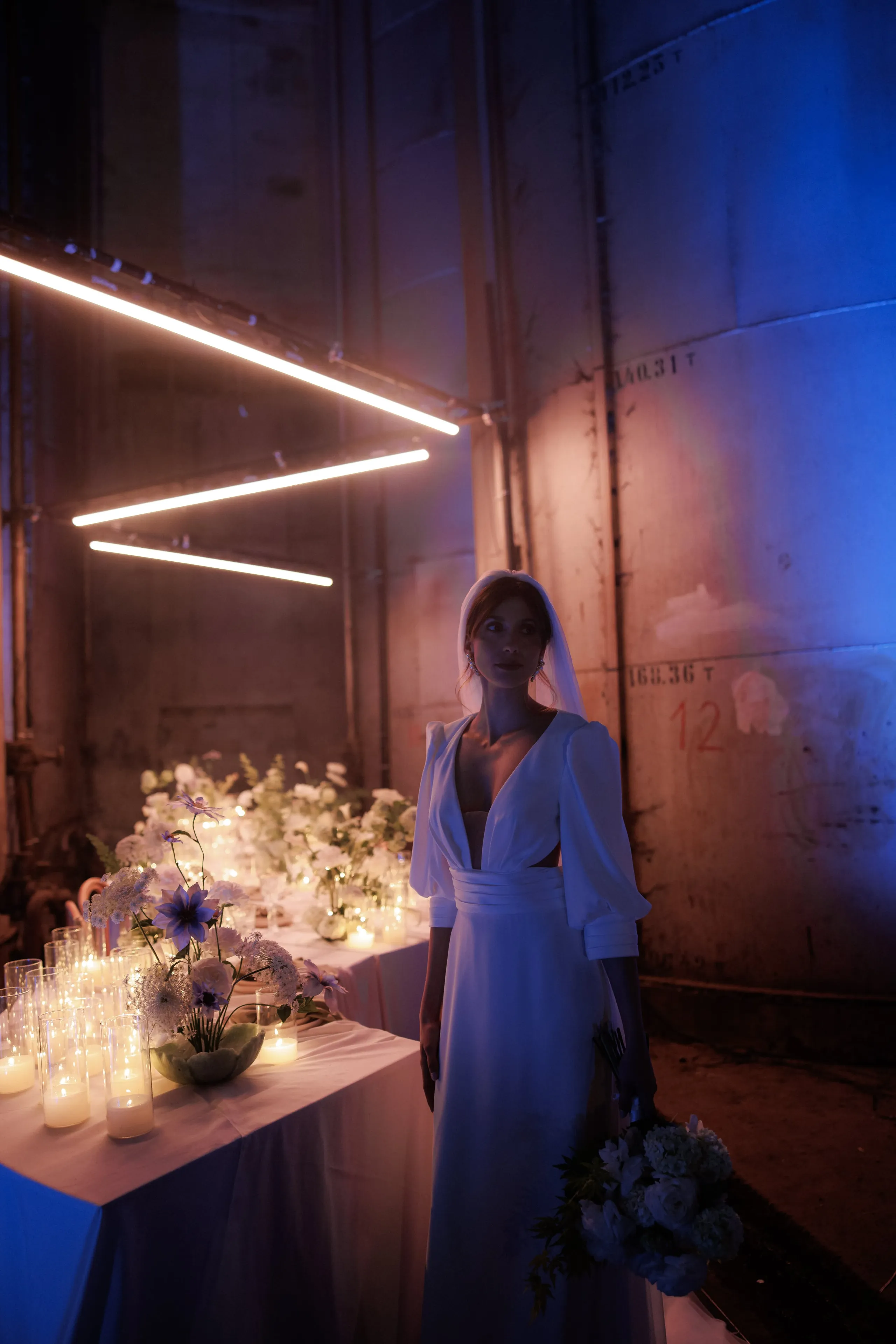 A bride in a white gown with a sheer veil stands in a dim industrial space in Rethimno, Crete, Greece, holding a loose bouquet of white roses and hydrangea-like blooms against her side. Behind her, long rows of wedding table flowers in white and pale blue, with purple clematis-style blooms, sit beside tall glass candle holders glowing amber. Blue and warm light washes over the concrete tanks, and neon-like linear lights zigzag overhead, creating a quiet, cinematic mood.