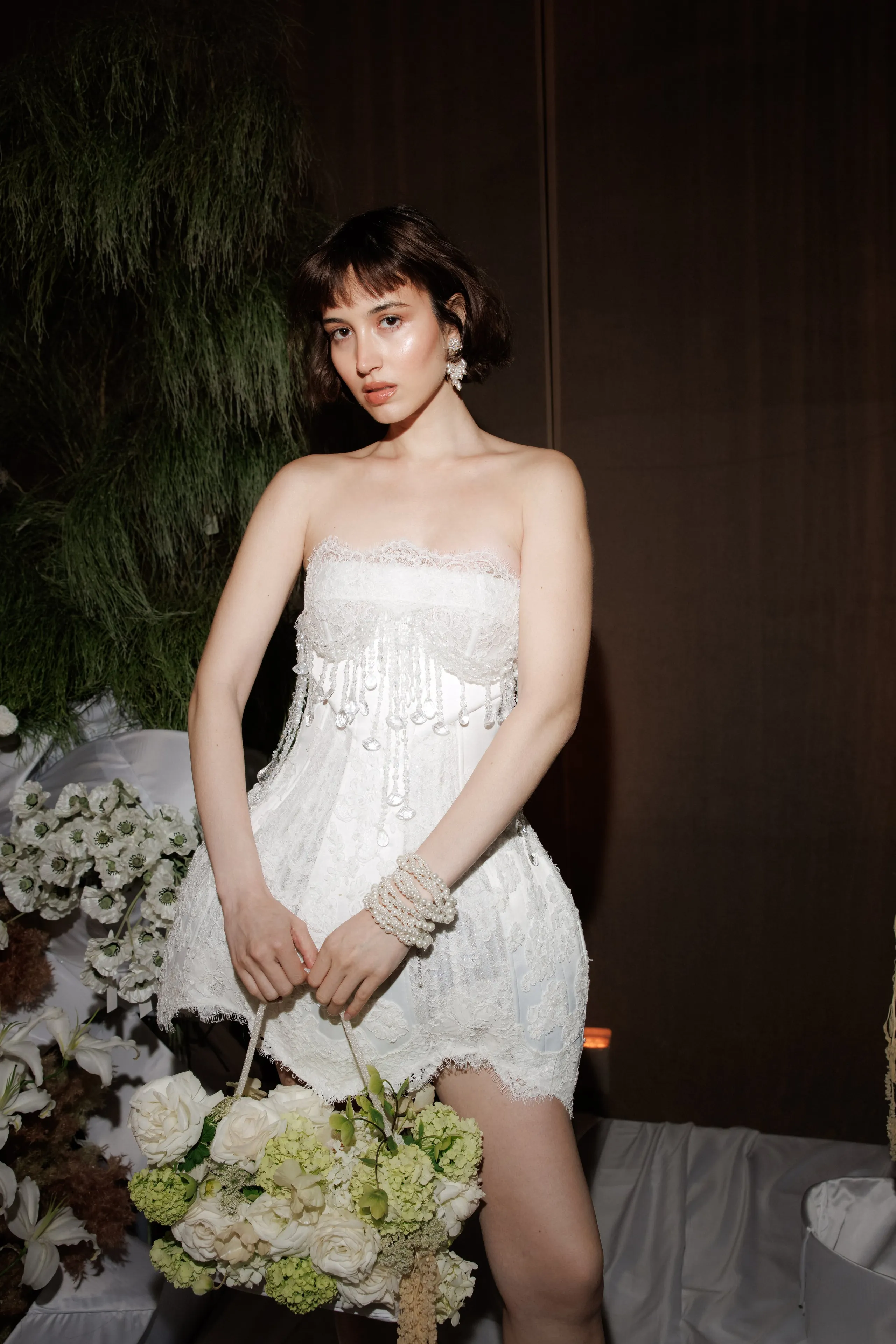 A young woman with a sleek dark bob stands in a short, strapless white lace dress trimmed with dangling crystal beads, holding a round bouquet of white garden roses and pale green hydrangeas by its handle. Behind her are a feathery green foliage backdrop and white flowers on a draped table, set against a dark wall in soft, low light. This is a wedding flower display from Rethimno, Crete, Greece, for the story The Extravaganza Flower.