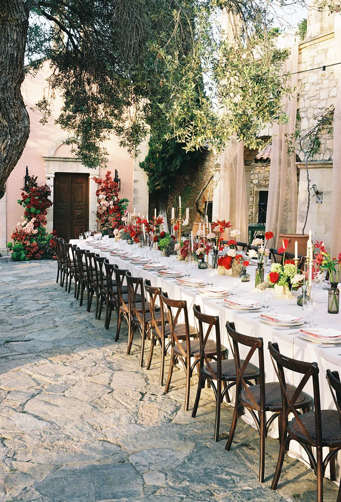 A long wedding dining table in a stone-paved courtyard at Agreco Farm in Greece, dressed with a white cloth, tall white candles, and centrepieces of red roses, pink garden roses, and green hydrangea in dark glass bottles. Tall arrangements of red blooms and green foliage stand beside a rustic wooden door in a pink-washed wall, and dark cross-back chairs line the table beneath olive branches in soft, warm afternoon light.