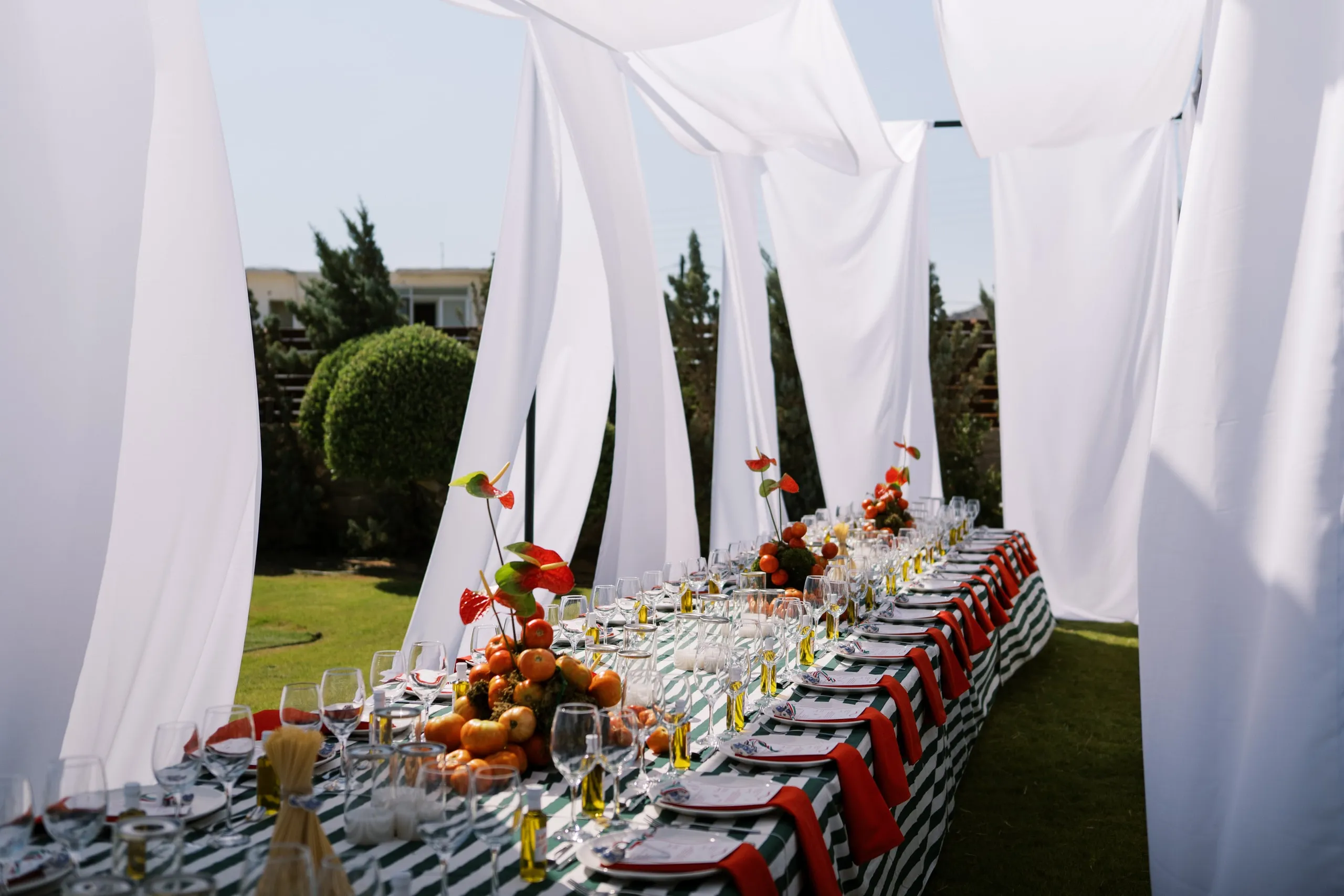 A long garden table dressed in a green and white striped cloth and red napkins, set with rows of wine glasses and white plates, runs beneath billowing white fabric drapes at Wine Dark Sea Villas in Rodia, Crete, Greece, where a wedding feast is laid out. Along its length sit low centrepieces of orange tomatoes and red anthurium blooms, with a few spires of the flowers rising above the fruit. Sheer white cloth hangs overhead in soft folds, and beyond it lie a manicured lawn, rounded green shrubs and a pale building under a clear sky.