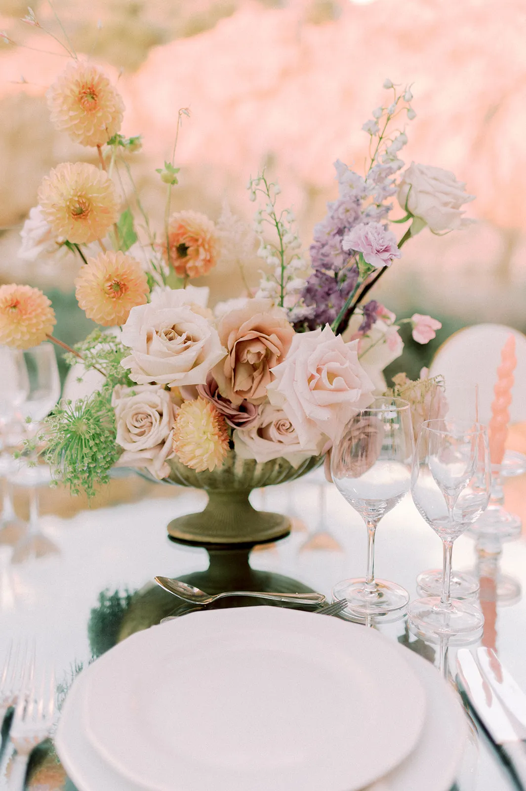 A wedding table centrepiece in a dark green footed urn, filled with soft blush and dusky peach garden roses, apricot dahlias, purple stock and green wild carrot. It sits on a white-clothed table beside clear stemless wine glasses and a silver spoon, with a pink taper candle at the edge, at Lake Vouliagmeni in Greece.