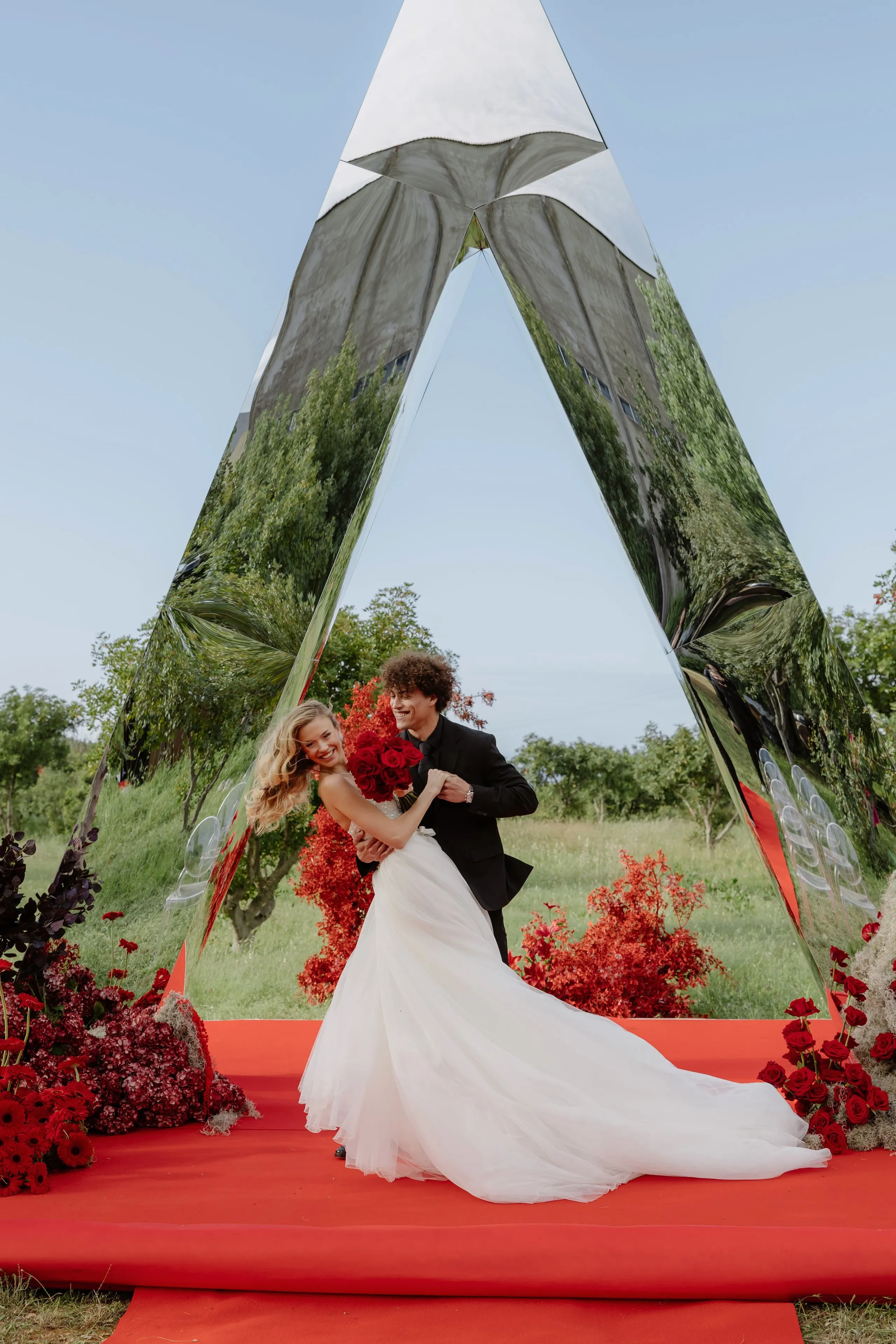 A bride in a flowing white gown with a long train stands on a red carpet, laughing over her shoulder, while a groom in a black suit holds her from behind and presses a red bouquet to her waist. They stand inside a tall, triangular mirrored arch that reflects the green trees and blue sky, and red flowers and foliage fill the space around them. This wedding flower display, with its striking red and white palette, is set in Rethimno, Crete, Greece.