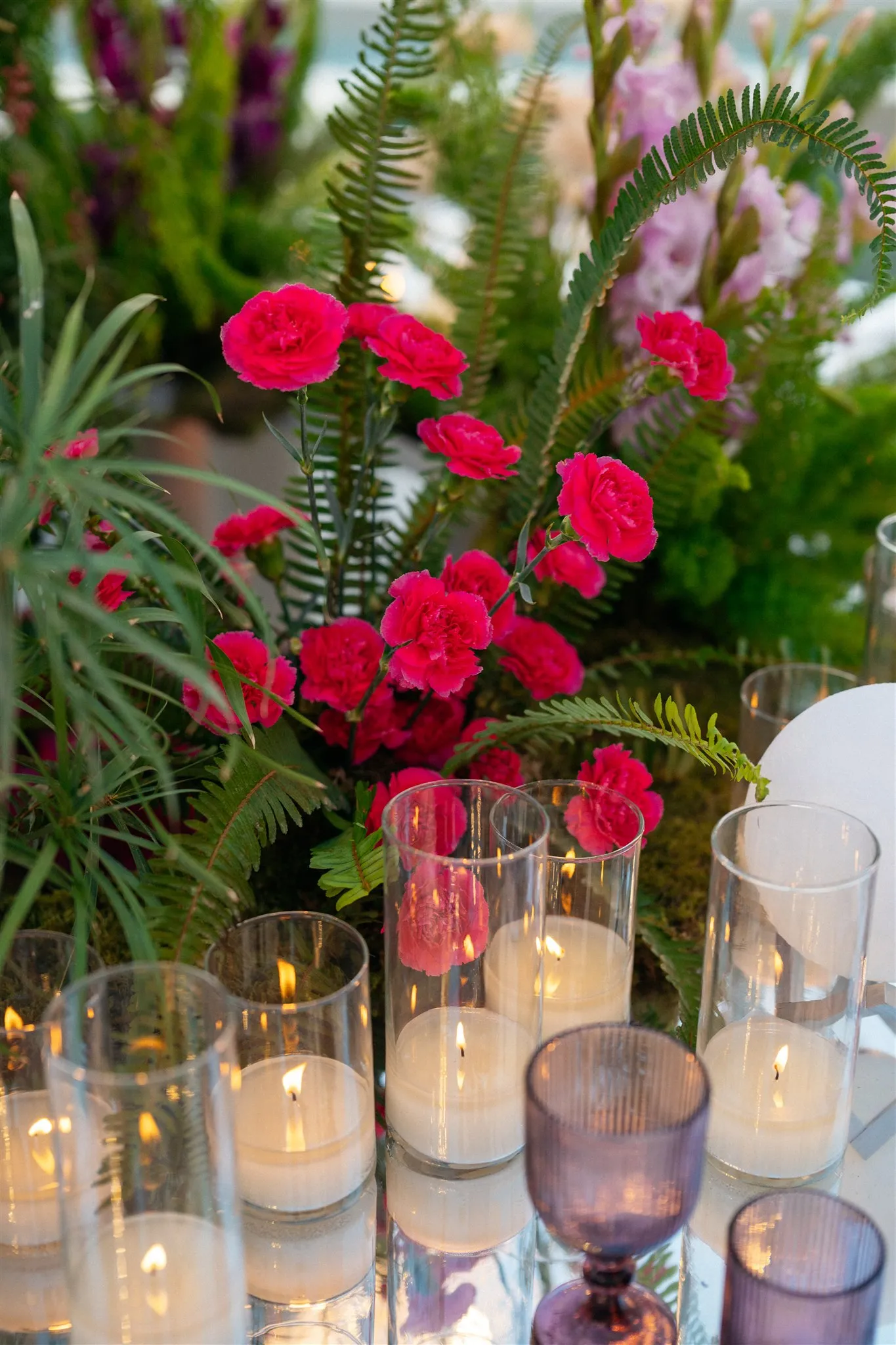 Vivid pink carnations rise from a lush cluster of feathery ferns and long green grass blades, with pale gladioli spiking up behind them, all arranged as a wedding table centrepiece at Islet in Thira, Santorini, Greece. Clear glass hurricane vases holding lit pillar candles stand in front of the blooms, their warm flames reflecting on the glossy table, while ribbed purple-glass goblets sit in the foreground.