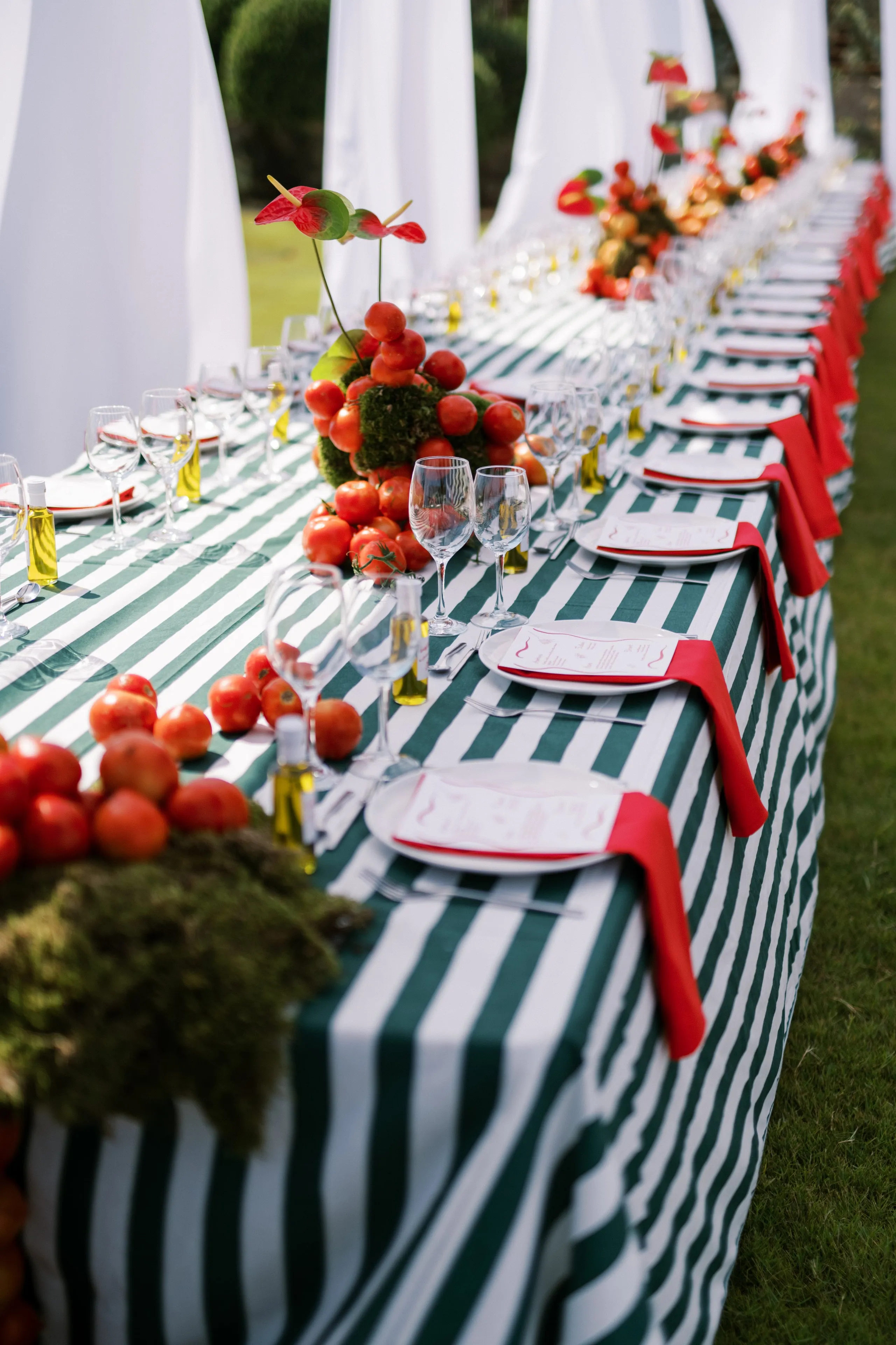 A long outdoor wedding table in Greece, set at Wine Dark Sea Villas in Rodia, Crete, draped in green and white striped linen with red napkins tucked beside each plate. Down the centre, low moss-covered domes are heaped with small red tomatoes, with red anthurium blooms rising from one, and clusters of orange and red flowers continue along the row. Stemmed glasses, small bottles of olive oil and white-tiled plates with red menu cards sit in neat rows beneath white draped canopies on a lawn.