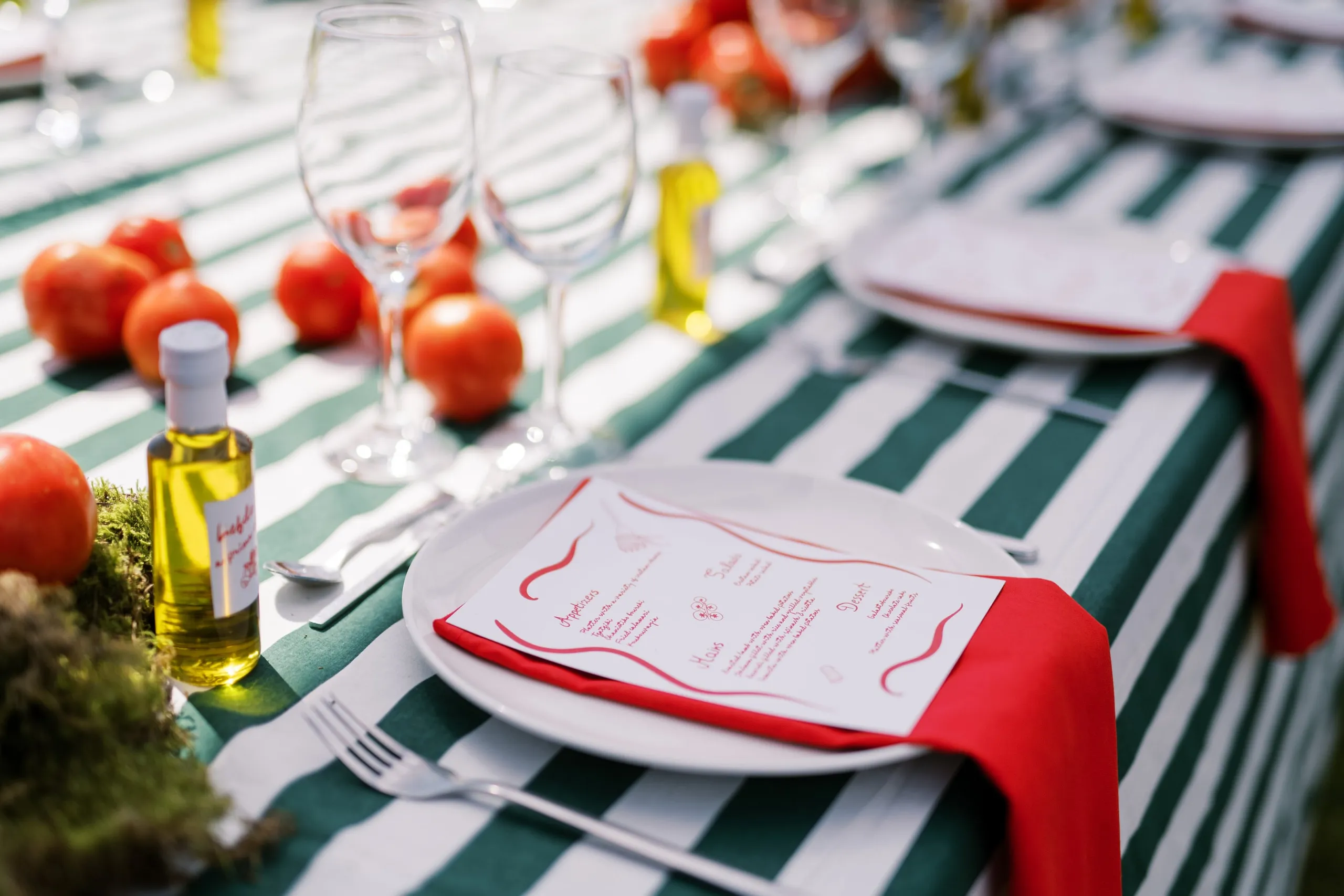 A long table dressed in green and white striped linen, set for a garden wedding dinner at Wine Dark Sea Villas on Crete in Greece. In the foreground a white plate holds a menu card with red hand-drawn swirls, tucked into a red napkin, with a silver fork beside it. Scattered across the table are ripe red tomatoes, small bottles of golden olive oil, clear wine glasses and a bed of green moss.