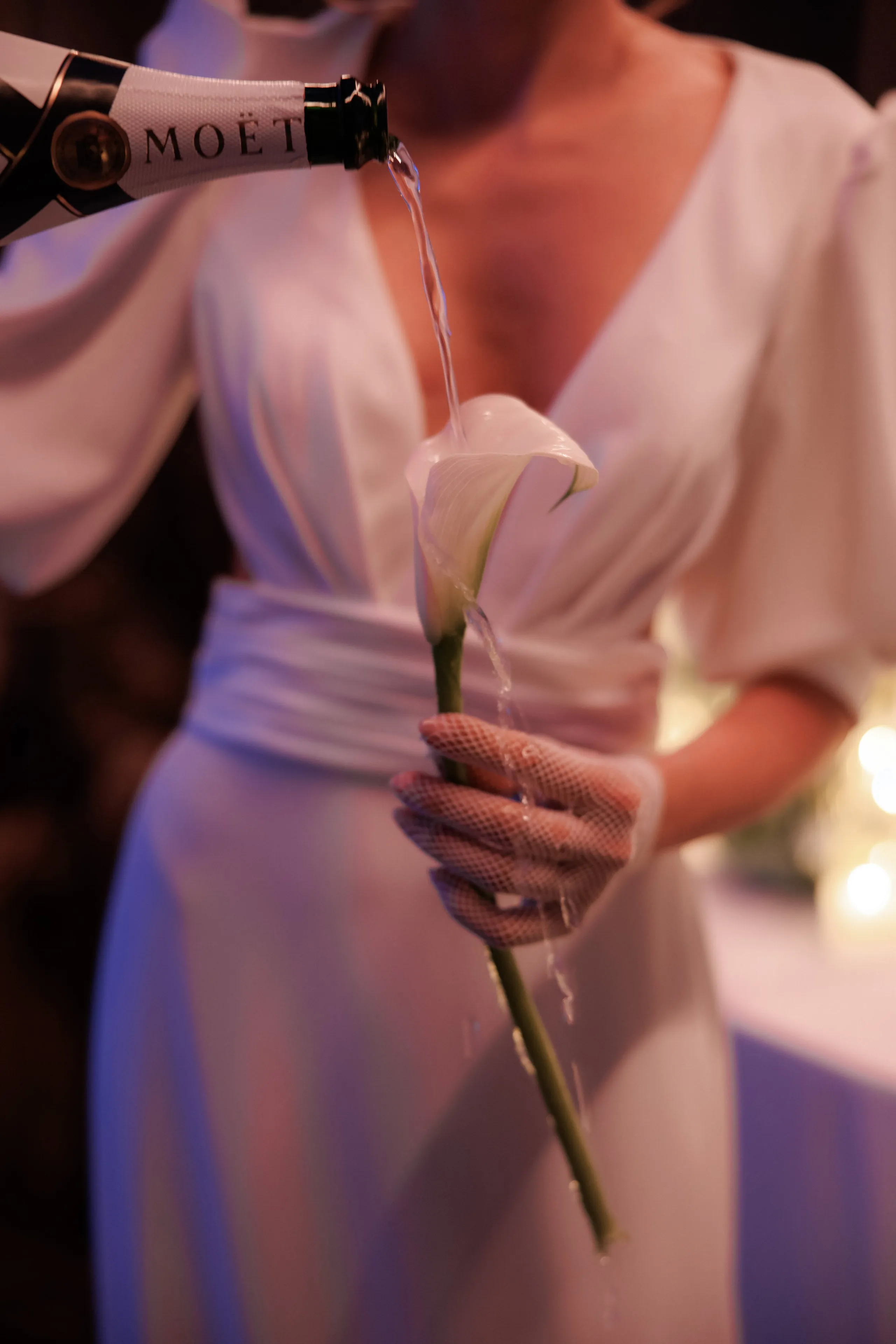 A close view of a single white calla lily held by a gloved hand in a white dress with a wide sash, as champagne from a Moët bottle is poured over the bloom and runs down its green stem. The soft, warm light and dark background give a glowing, intimate mood at this wedding flower moment in Rethimno, Crete, Greece.