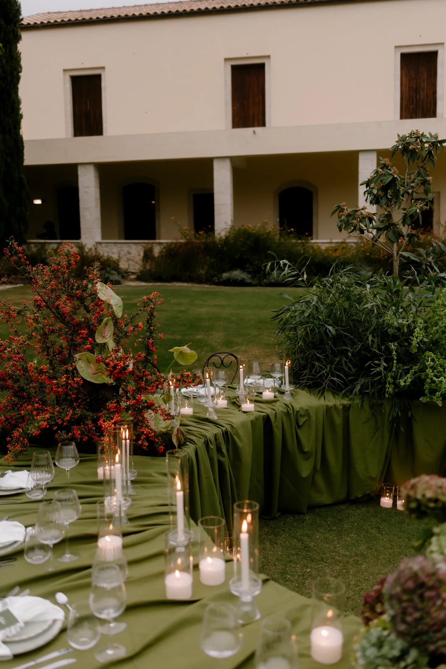 Long wedding dining tables draped in deep olive-green linen run across a lawn at Ktima Roxani Estate in Crete, Greece, lit by rows of pillar and taper candles in glass hurricanes. At left, a tall arrangement of red berried branches is dotted with green anthurium leaves, and at right a billowing mass of slender green foliage softens the table's edge. Behind them, a cream two-storey estate building with arched openings and wooden shutters glows in the dusk.