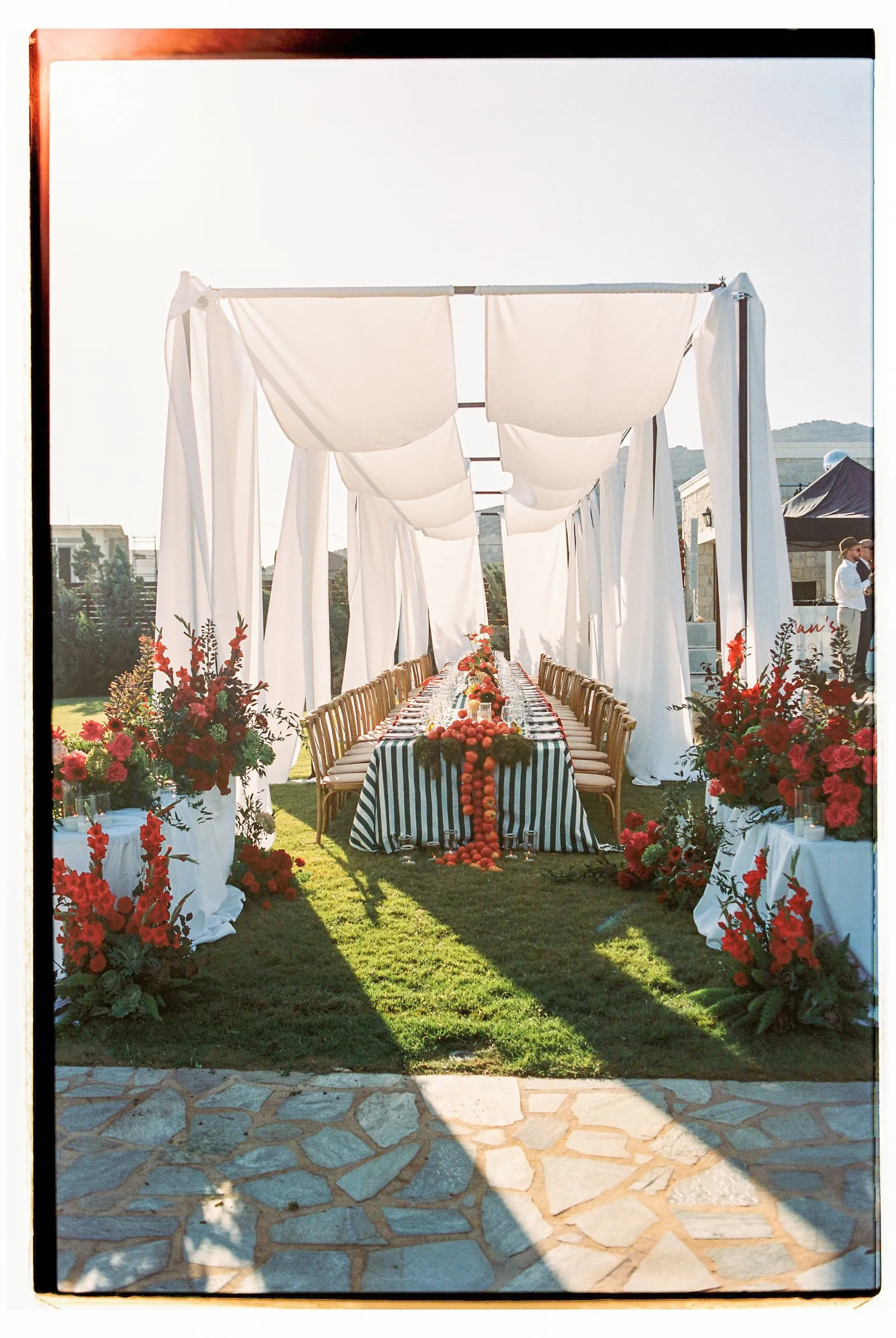 A long garden dining table set for a wedding at Wine Dark Sea Villas in Rodia, Crete, Greece, draped beneath billowing white fabric hung from a pale frame. The table has a navy and white striped runner and is lined with wooden chairs, with a trailing garland of orange-red blooms and deep green foliage running down its centre. Tall arrangements of red gladioli and pink-red roses flank the scene on white-draped stands, and two people in white stand in the distance at the right, in warm late-afternoon sunlight.