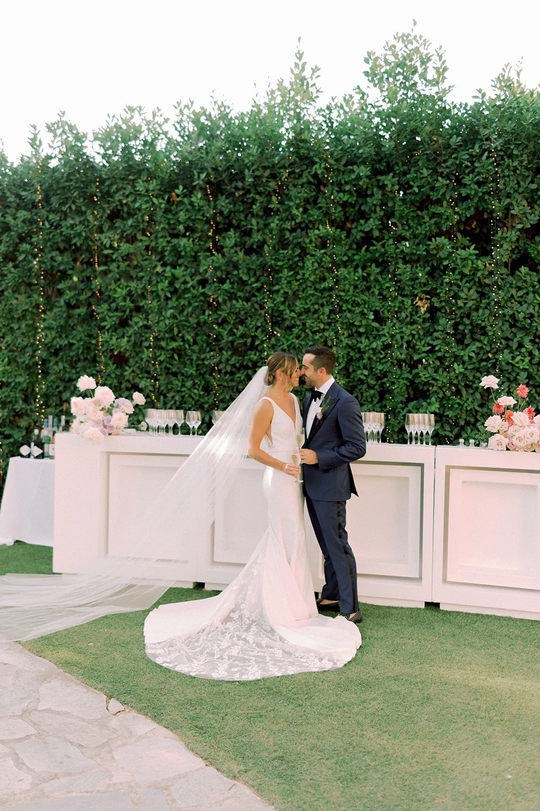 A bride in a fitted white gown with a long cathedral veil and a lace-patterned train stands close to her groom in a navy tuxedo with a white rose boutonniere, the two leaning in nose to nose. They are beside a white bar decorated with soft pink and blush garden roses, wine glasses in rows, and a dense green hedge strung with fairy lights, in a wedding flower setting at Lake Vouliagmeni, Greece.