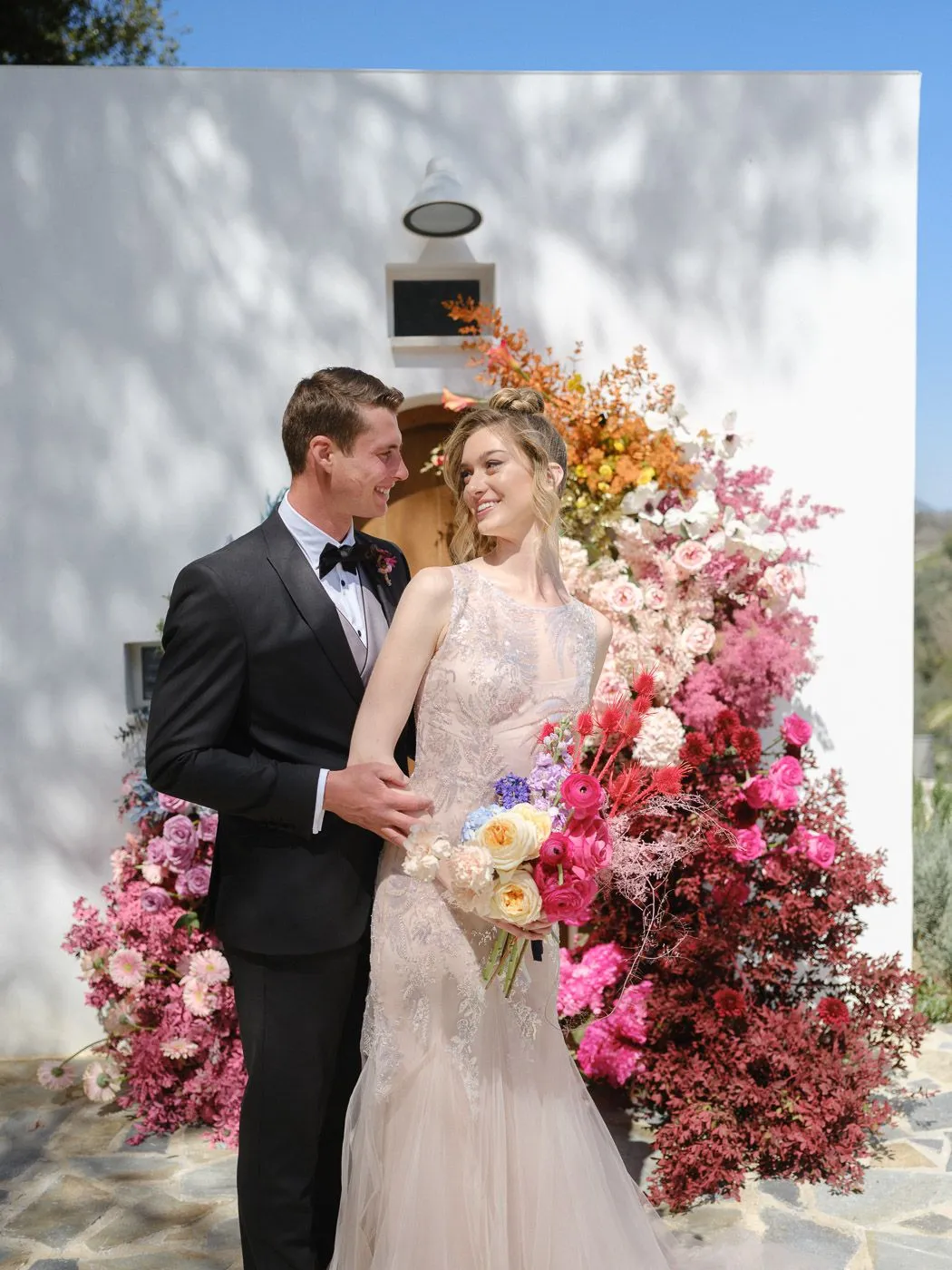 A bride and groom smile at each other in front of a whitewashed wall covered in a dramatic floral installation that fades from orange and gold at the top to blush, hot pink and deep berry red below. The bride wears a fitted blush gown with delicate embroidery and holds a bouquet of cream roses, pink ranunculus and red thistle-like blooms, while the groom wears a black tuxedo and bow tie. This wedding flower display at Markakis Estate in Emprosneros, Crete, Greece, is set on a stone terrace under bright sunshine.