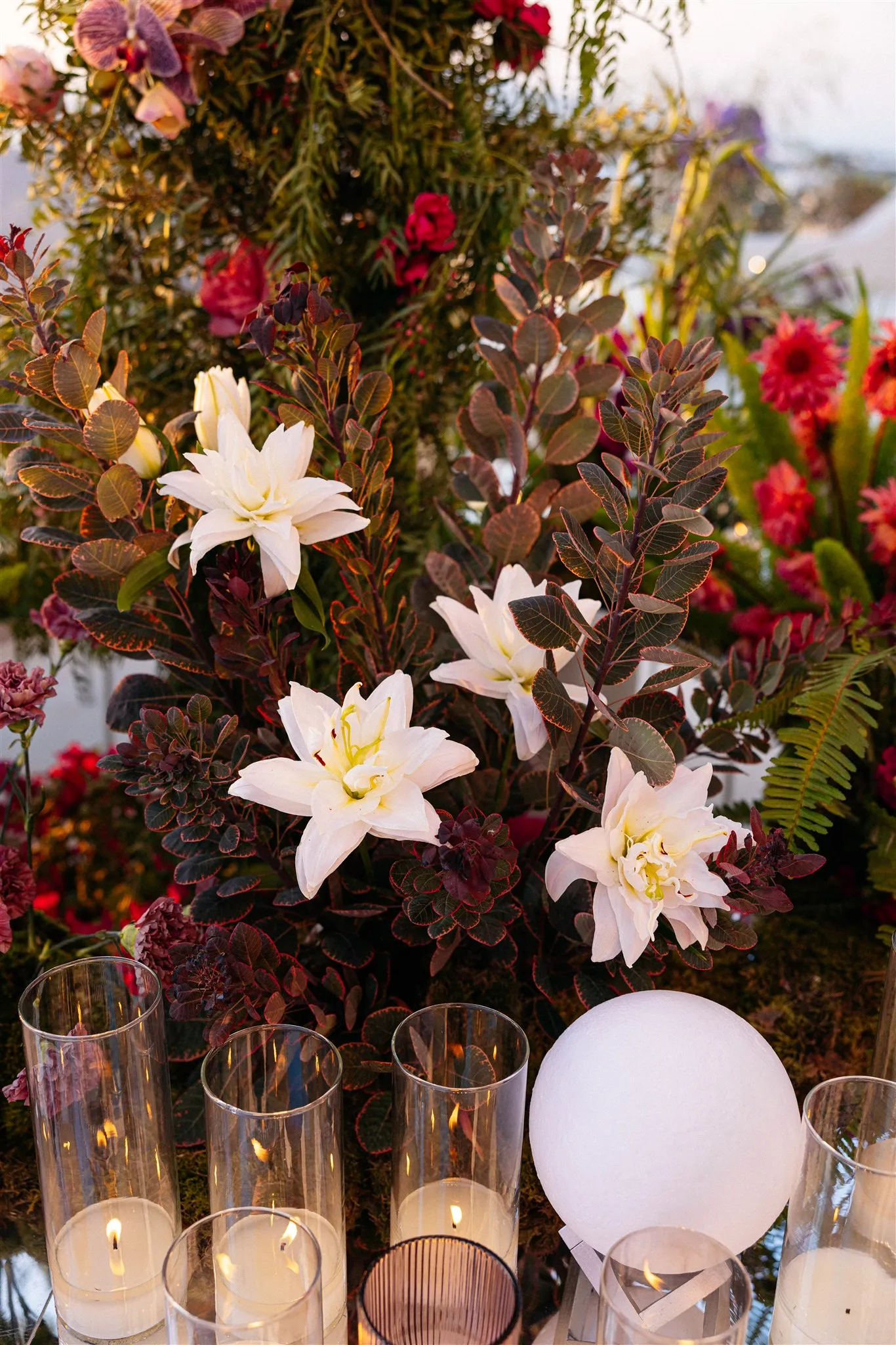 Close-up of a lush table arrangement at Islet Santorini in Thira, Greece, for a wedding: creamy white double lilies rise from a bed of dark burgundy foliage, with deep red roses and ferns tucked behind. In the foreground, several clear glass cylinders hold glowing pillar candles, alongside a smooth white sphere, all set on moss. The warm evening light and rich, moody colours give the flowers a dramatic, candlelit feel.