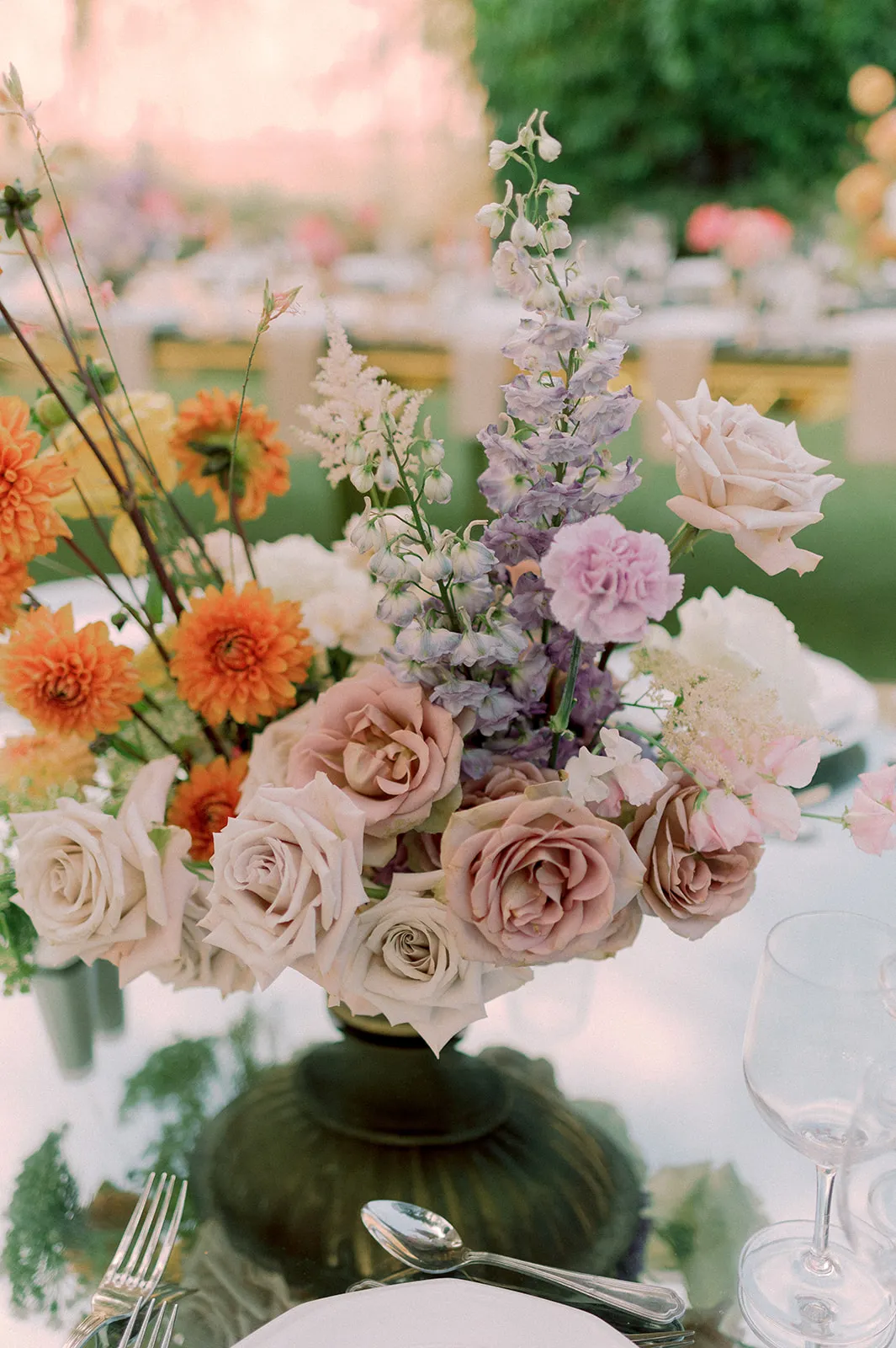 A tall centrepiece on a wedding table at Lake Vouliagmeni in Greece, built around dusky blush and mauve garden roses in a dark green footed vase. Above them rise orange dahlias, lilac delphinium, a pale pink carnation and feathery astilbe, with a few white sweet peas trailing at the side. Soft daylight falls on the white tablecloth, and a stemmed wine glass and a silver fork sit at the edge of the frame.