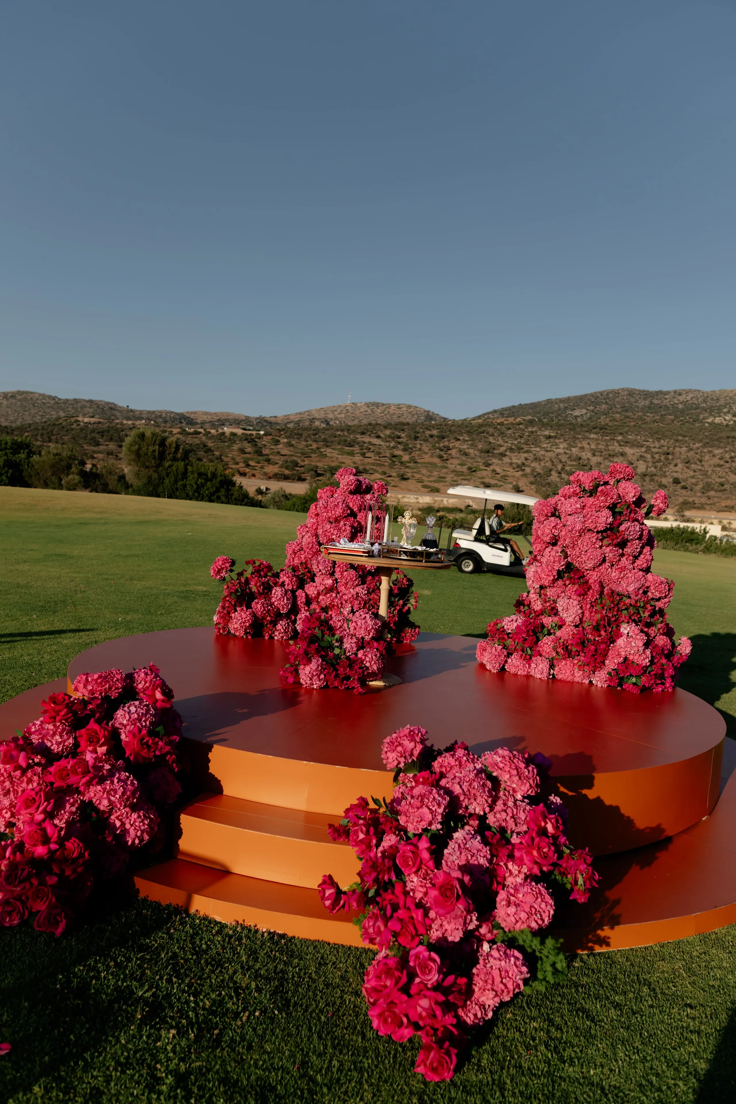 A circular terracotta-orange stage stands on a lush green lawn, its stepped edges framed by dense clusters of pink and fuchsia flowers, including hydrangeas and garden roses, with a tall cluster rising at each side. Wedding flowers at Crete Golf Club in Chersonisos, Greece, fill the platform with saturated pink, while a small gilded table with a few ornaments sits among the blooms. Beyond, dry brown hills and a clear blue sky set a calm, sunlit mood, with a golf buggy passing in the distance.