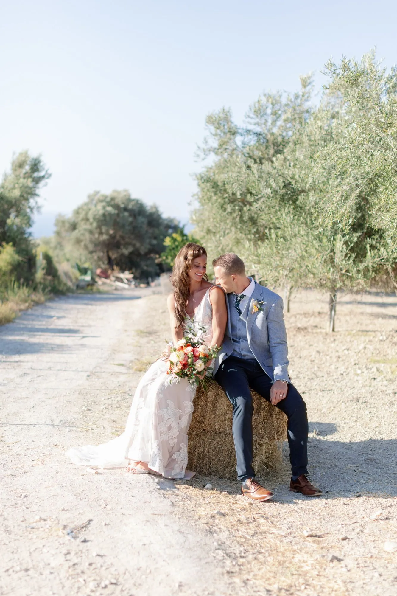 A bride in a lace-appliquéd gown sits on a round hay bale beside a dusty gravel track, holding a loose bouquet of coral and cream roses with greenery, while a groom in a light grey suit and navy trousers leans in close to her. Tall olive trees line the path under a pale, sunlit sky, and the soft morning light gives the scene a calm, romantic mood. This wedding portrait was taken at AgrecoFarms in Adele, Crete, Greece.
