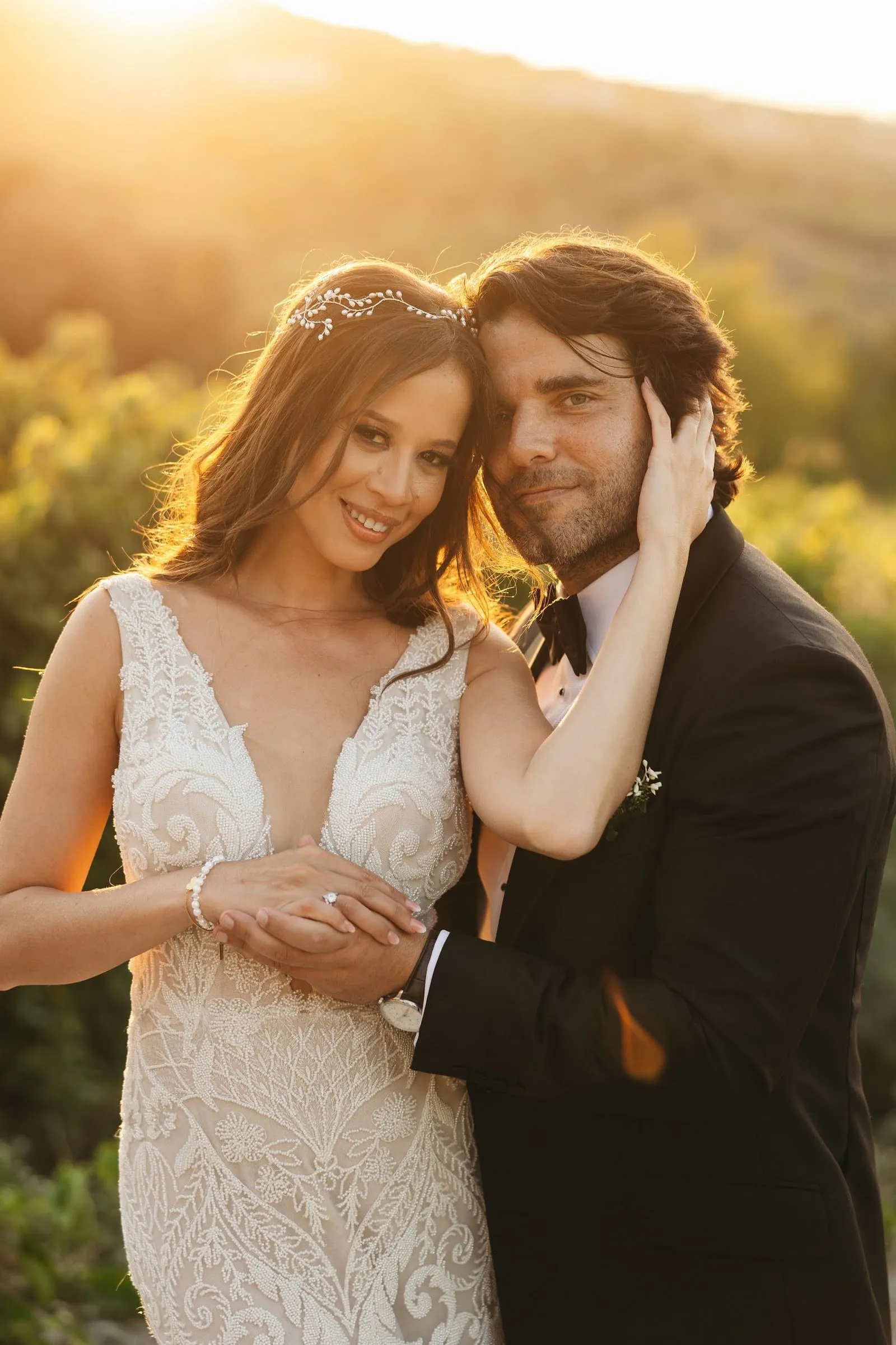 A bride and groom embrace in warm golden sunset light at AgrecoFarms in Adele, Crete, Greece. The bride wears a fitted, ivory beaded gown with a deep V neckline and a delicate pearl-and-crystal vine in her hair, while her hand rests over the groom's. He wears a black tuxedo with a small white boutonniere and leans in close, his hand on her cheek. Soft-focus green foliage and rolling hills glow behind them.