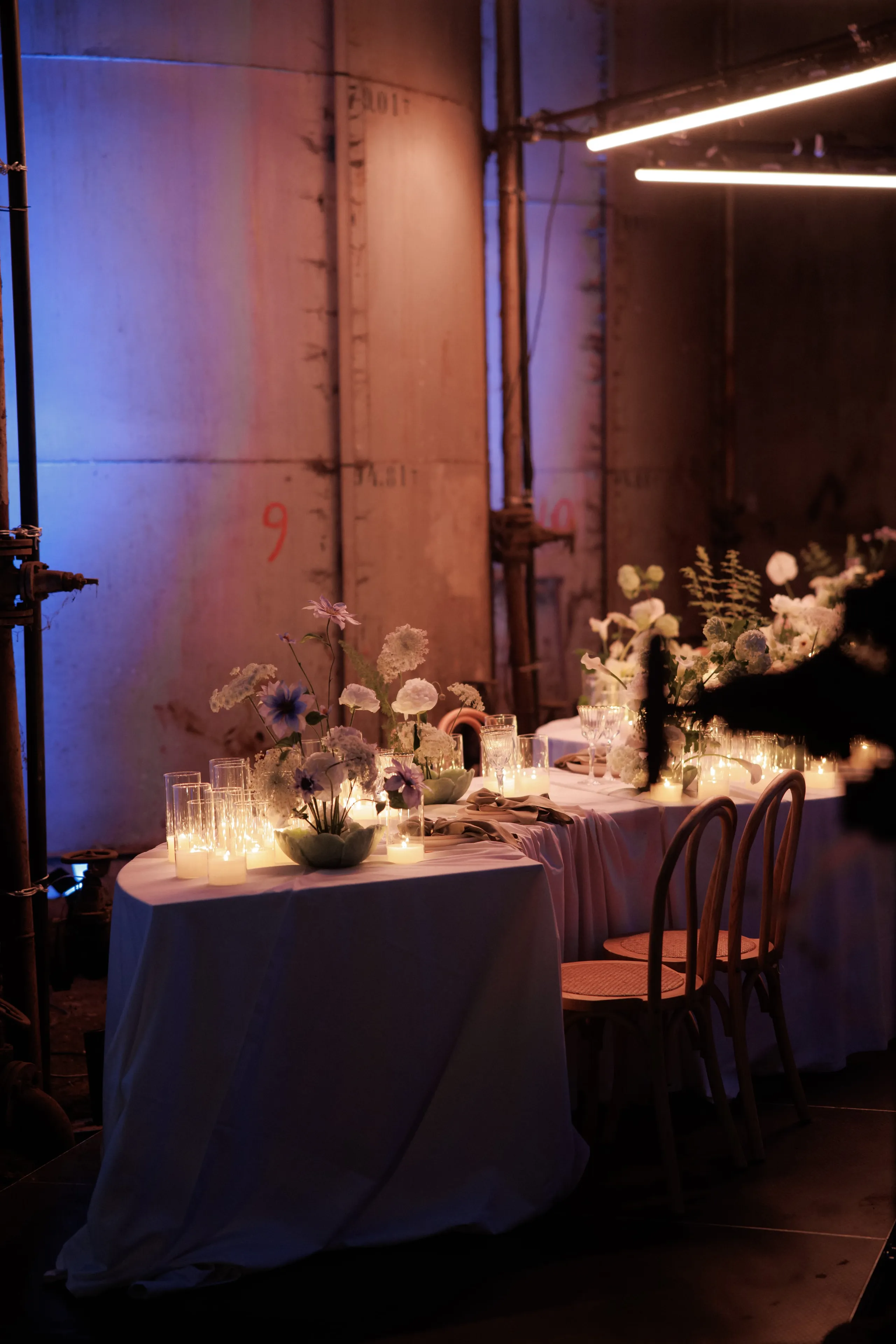 Two round-cornered tables draped in white linen sit in a dim industrial hall at Rethimno, Crete, Greece, with weathered concrete walls and bare metal scaffolding behind them. The low centrepieces hold pale blue and lilac flowers, white hydrangea and ranunculus-style blooms in soft green bowls, surrounded by clusters of glass votive candles whose warm light glows against the cool violet wall. Empty bentwood chairs with cane seats wait along the table, and a linear light fixture overhead adds a clean line to the scene. This is a wedding table styling from Greece.