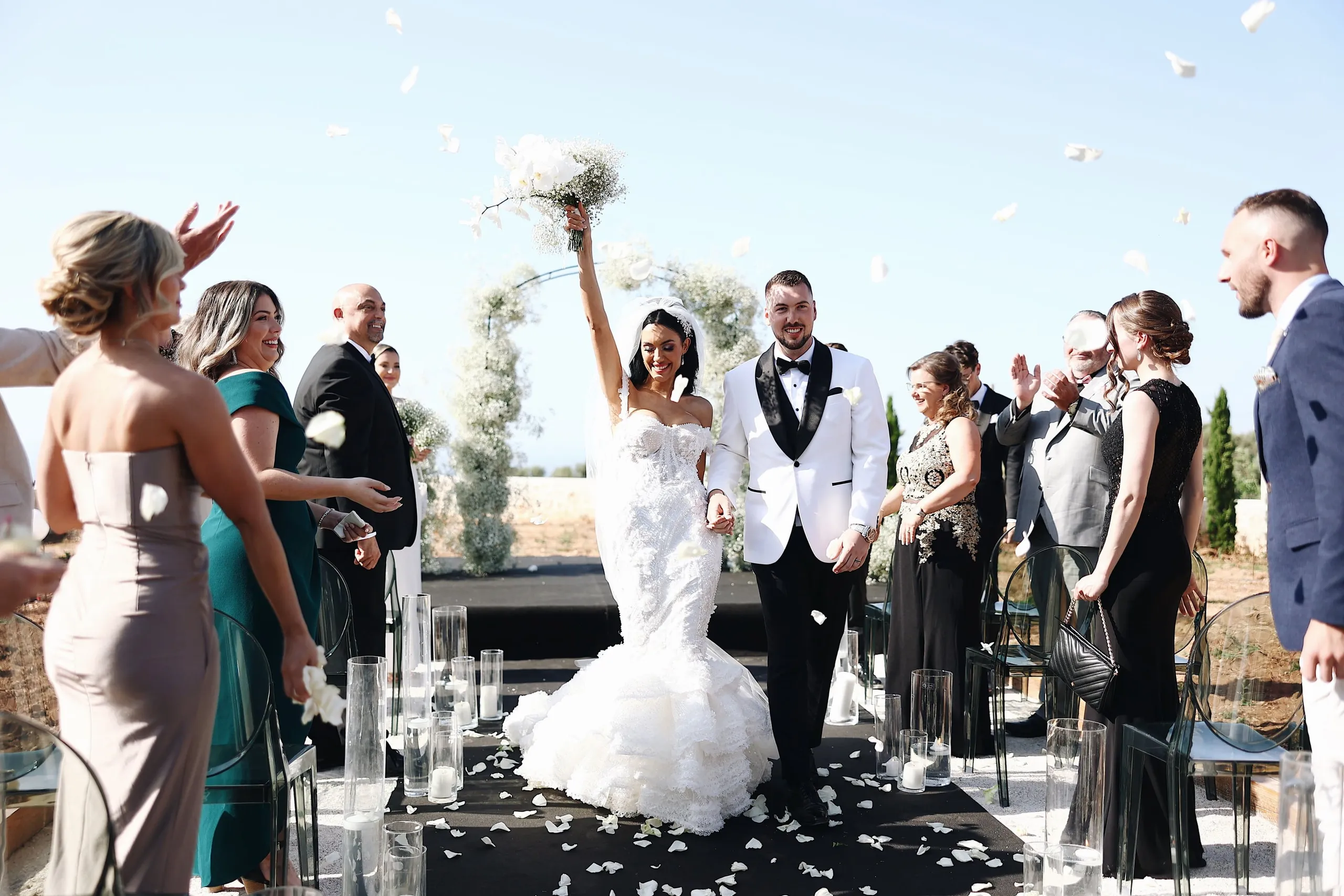 A bride in a fitted, lace-covered mermaid gown raises a white-and-cream bouquet of roses and baby's breath high in one hand as she walks down a black aisle at Villa Amnalys in Amnatos, Crete, Greece, beside her smiling groom in a white dinner jacket with black lapels. White petals drift through the bright sky and scatter across the dark runner, while guests on either side clap and cheer from clear acrylic chairs lined with candle-filled glass hurricanes. Behind them stands a white-flowered arch of baby's breath and blooms.