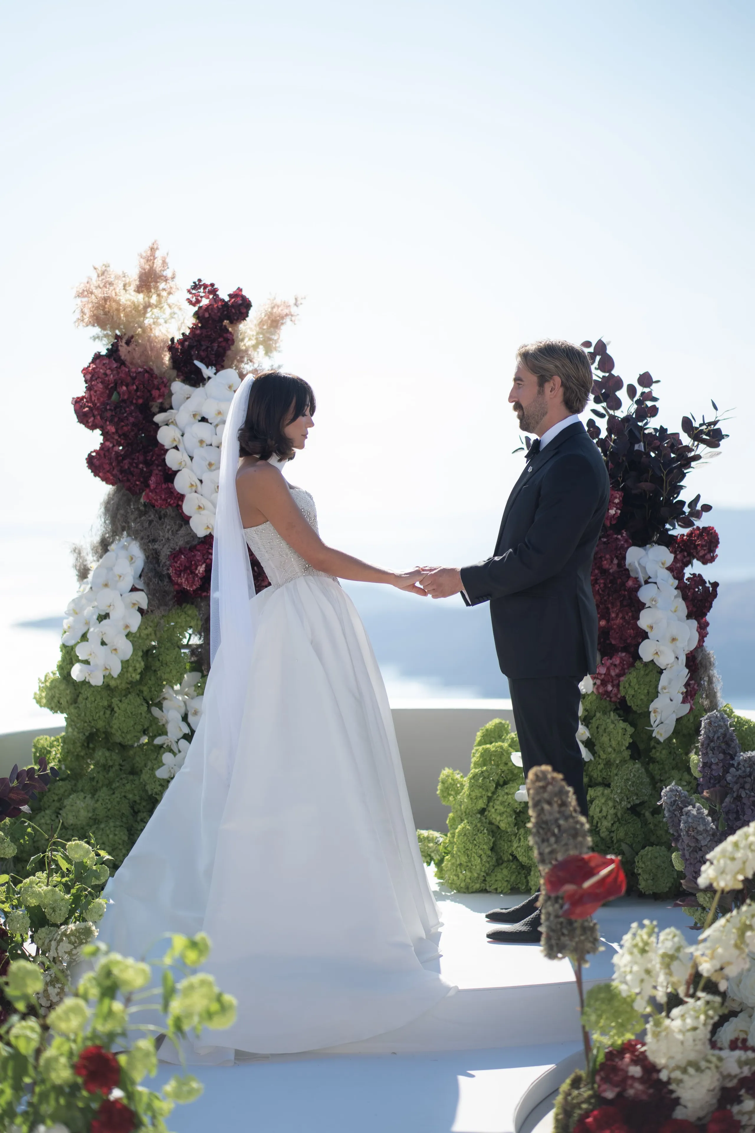 A bride in a white strapless gown with a long veil and a groom in a black suit and bow tie hold hands and face each other on a white platform at Islet Santorini in Thira, Greece. Behind them an arch of white orchids, deep burgundy hydrangeas, dark eggplant foliage, and green hydrangea clusters frames a pale, hazy sky over the sea. Red anthurium and white blooms in the foreground complete this wedding floral design in Greece.